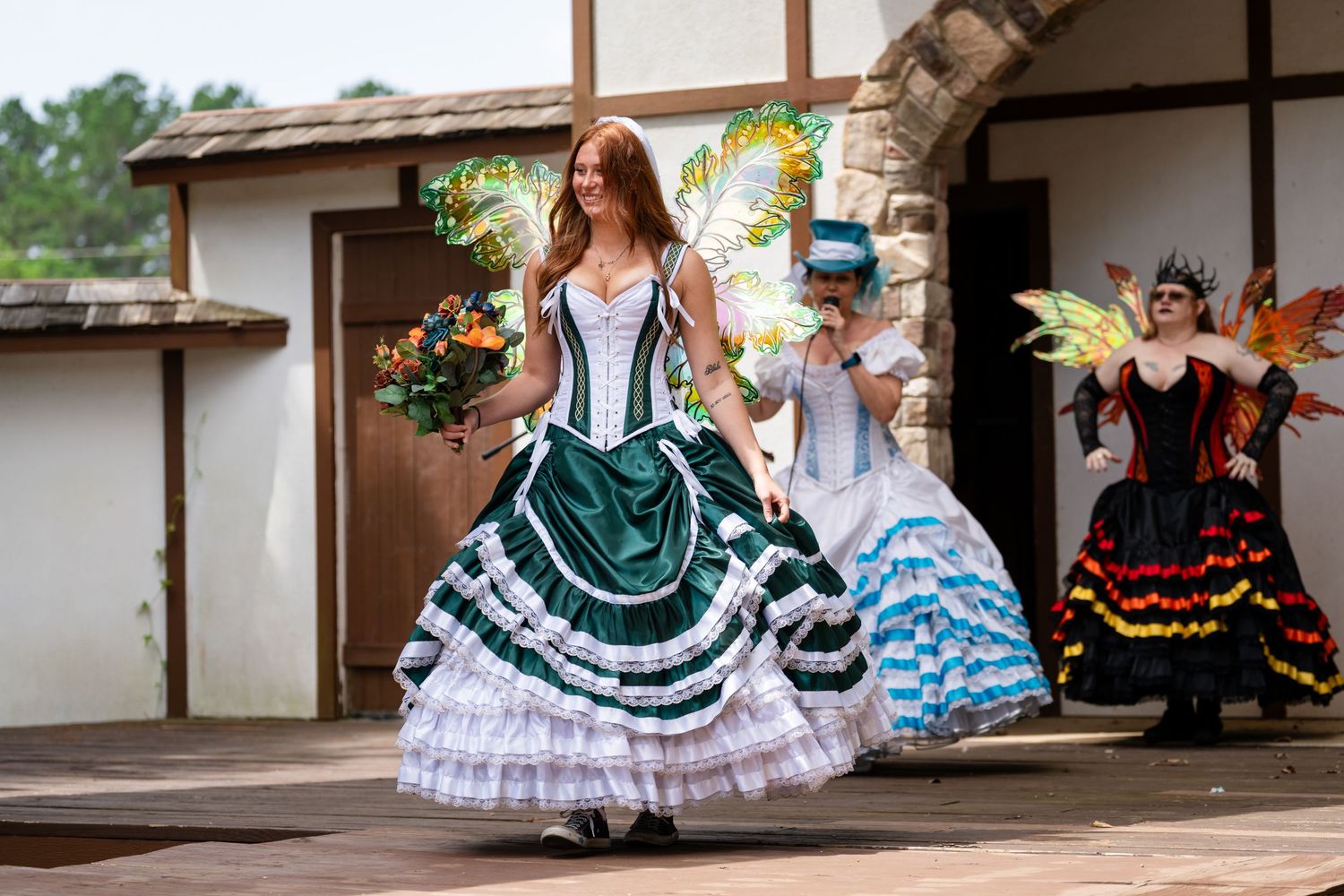 Three women in elaborate fairy tale ball gowns with colorful wings perform at an outdoor Renaissance festival stage.