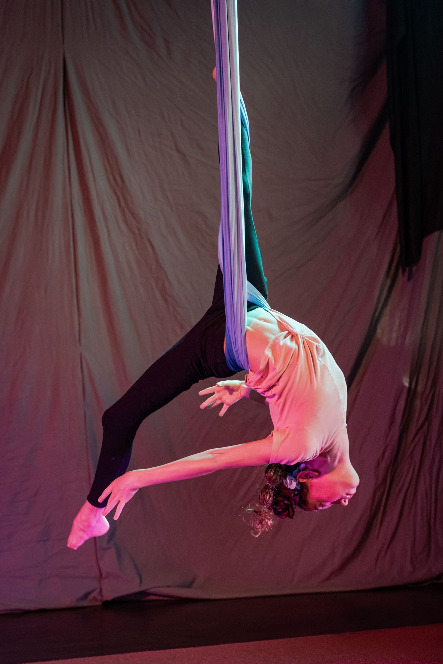 Aerial silk performer inverted in graceful backbend pose, wrapped in purple fabric against draped backdrop.