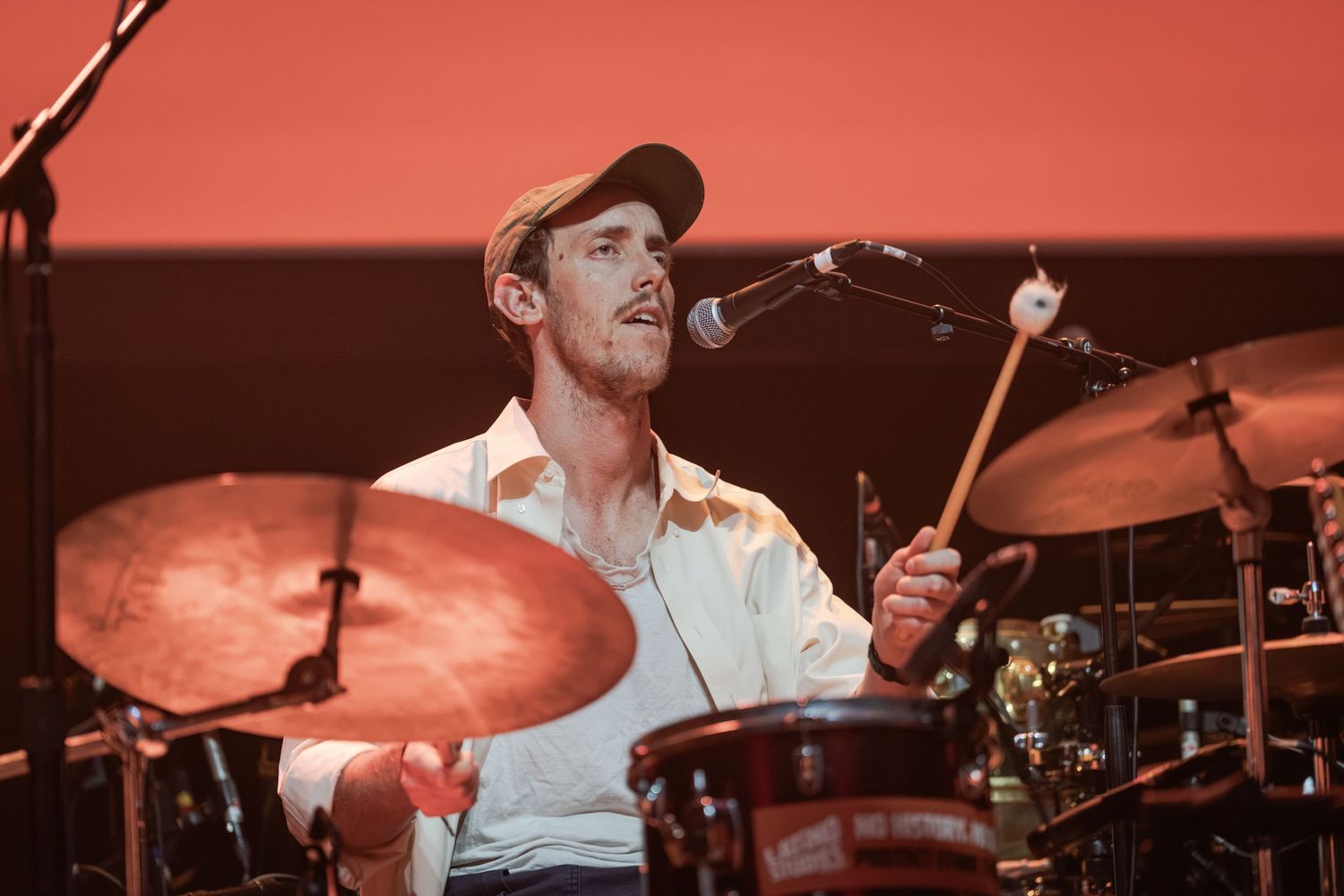Male drummer playing cymbals on stage with microphone, wearing cap and white shirt against red backdrop.