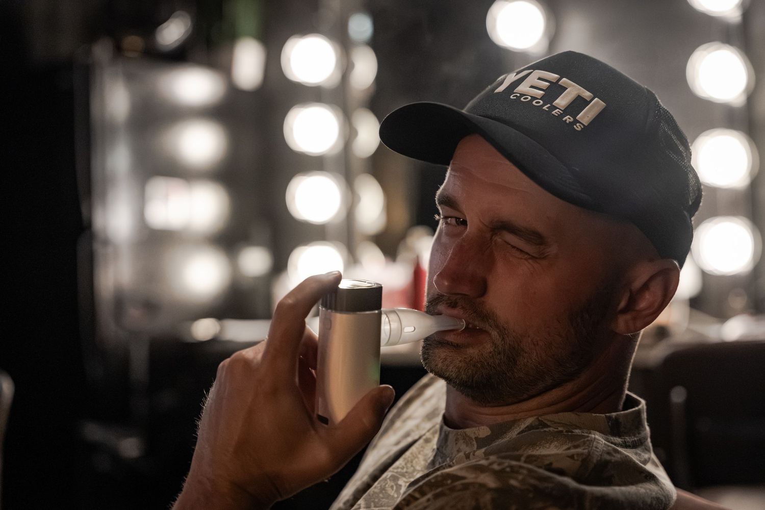 Man wearing a YETI cap drinking from a tumbler in a dimly lit room with bright vanity lights in the background.