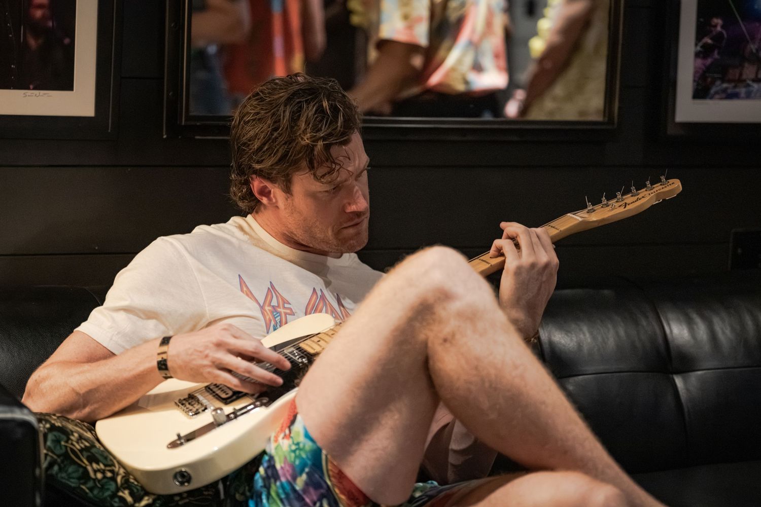 Man in colorful shorts and white tee plays electric guitar while sitting on a couch in a music room.