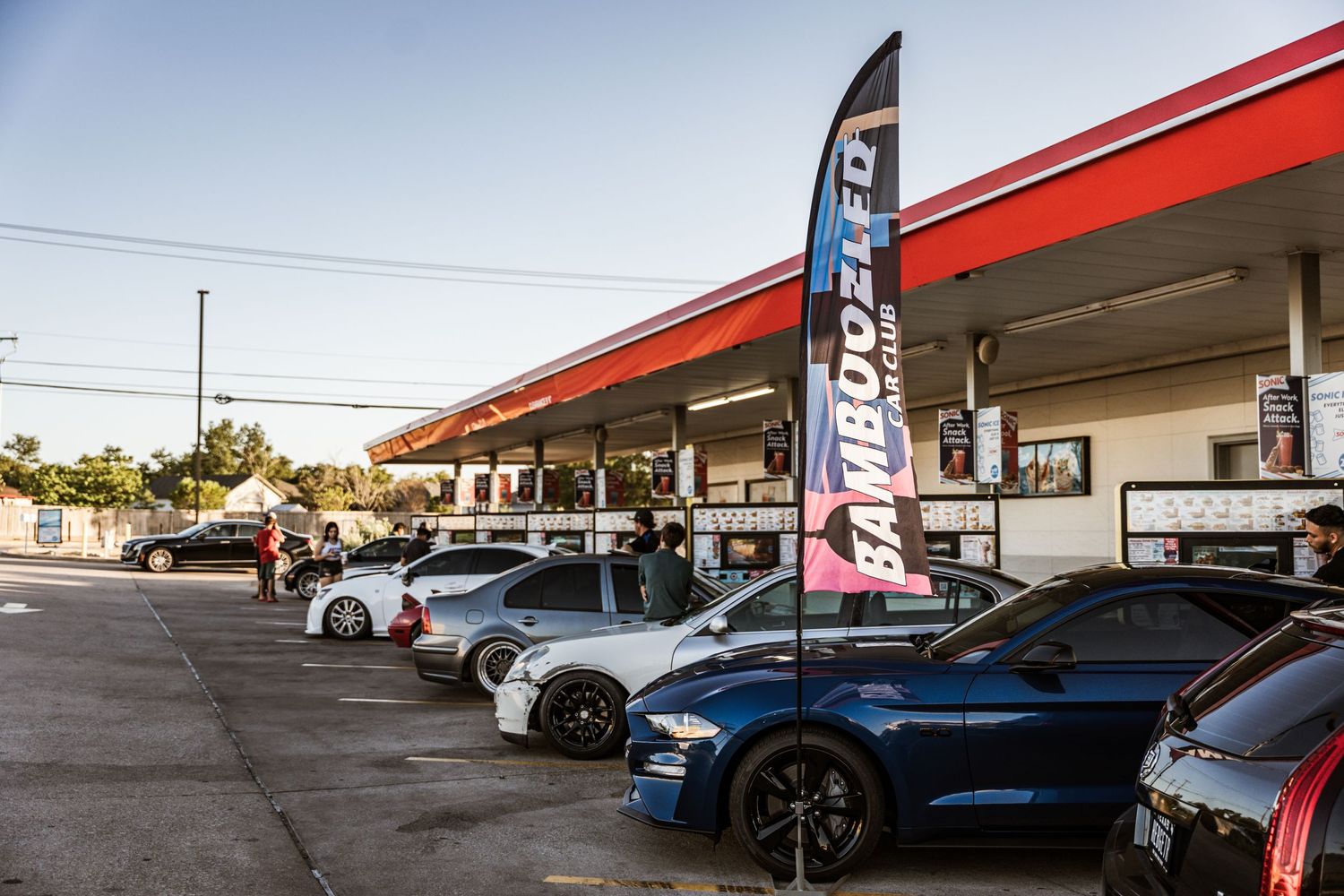 Row of modified cars parked at gas station with American Muscle feather flag.