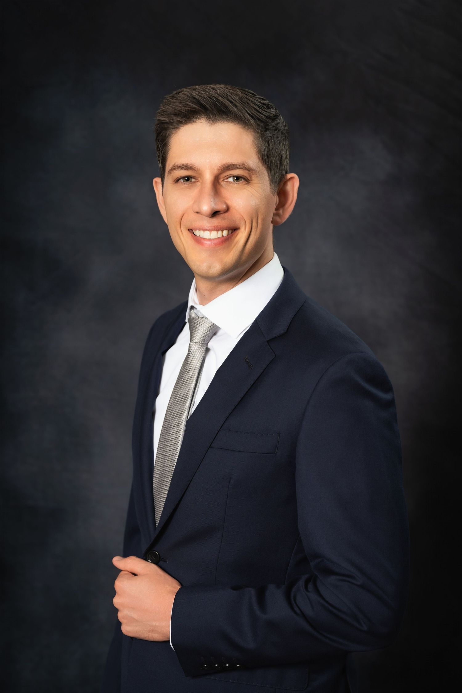 Professional headshot of a businessman in a navy suit and gray tie against a dark background.
