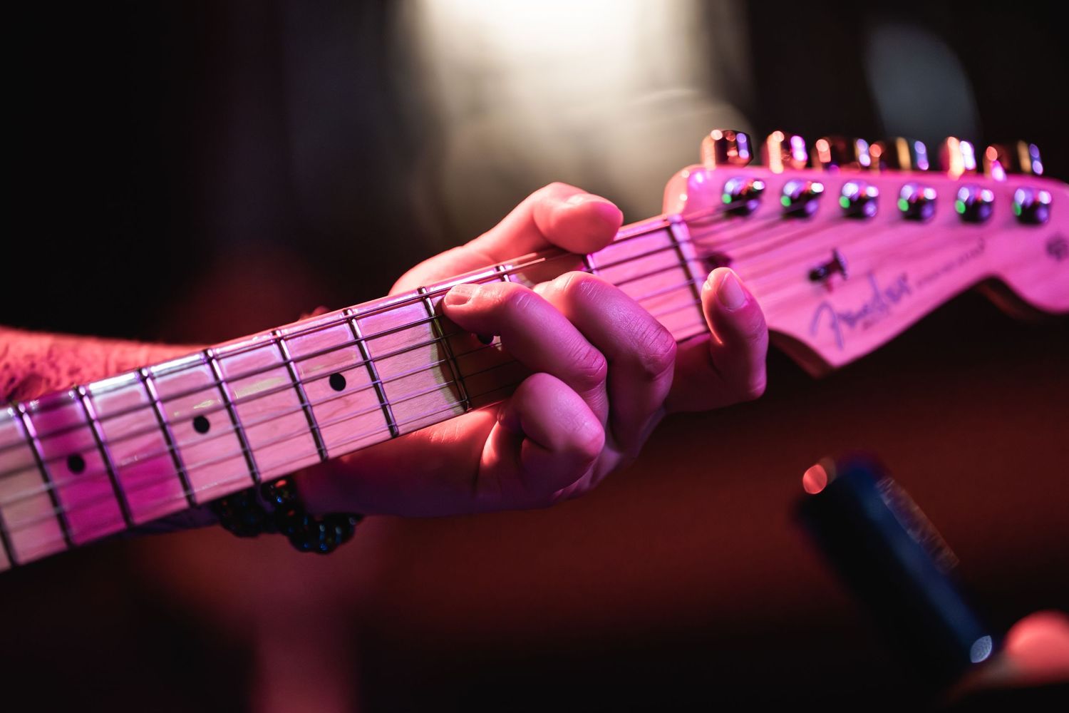 Close-up of hand playing pink-illuminated Fender guitar fretboard.