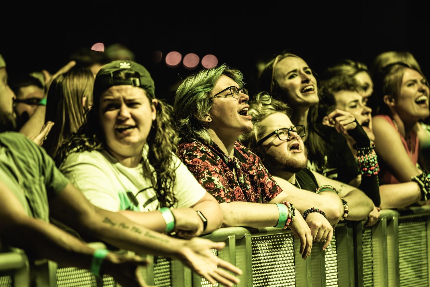 Concert crowd leans eagerly against barricade under yellow-green lighting during live music show.