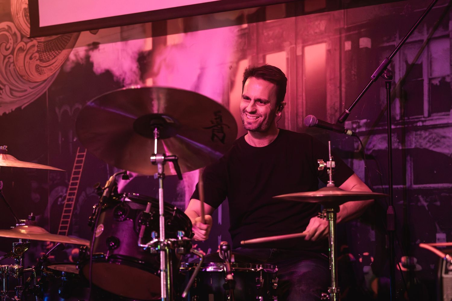 Drummer performs on stage bathed in pink and purple lighting at a live music venue.