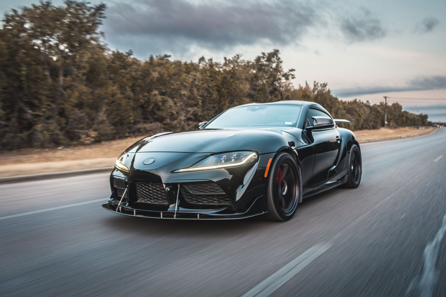 Black Toyota Supra with widebody kit and red accents speeding down a scenic road.