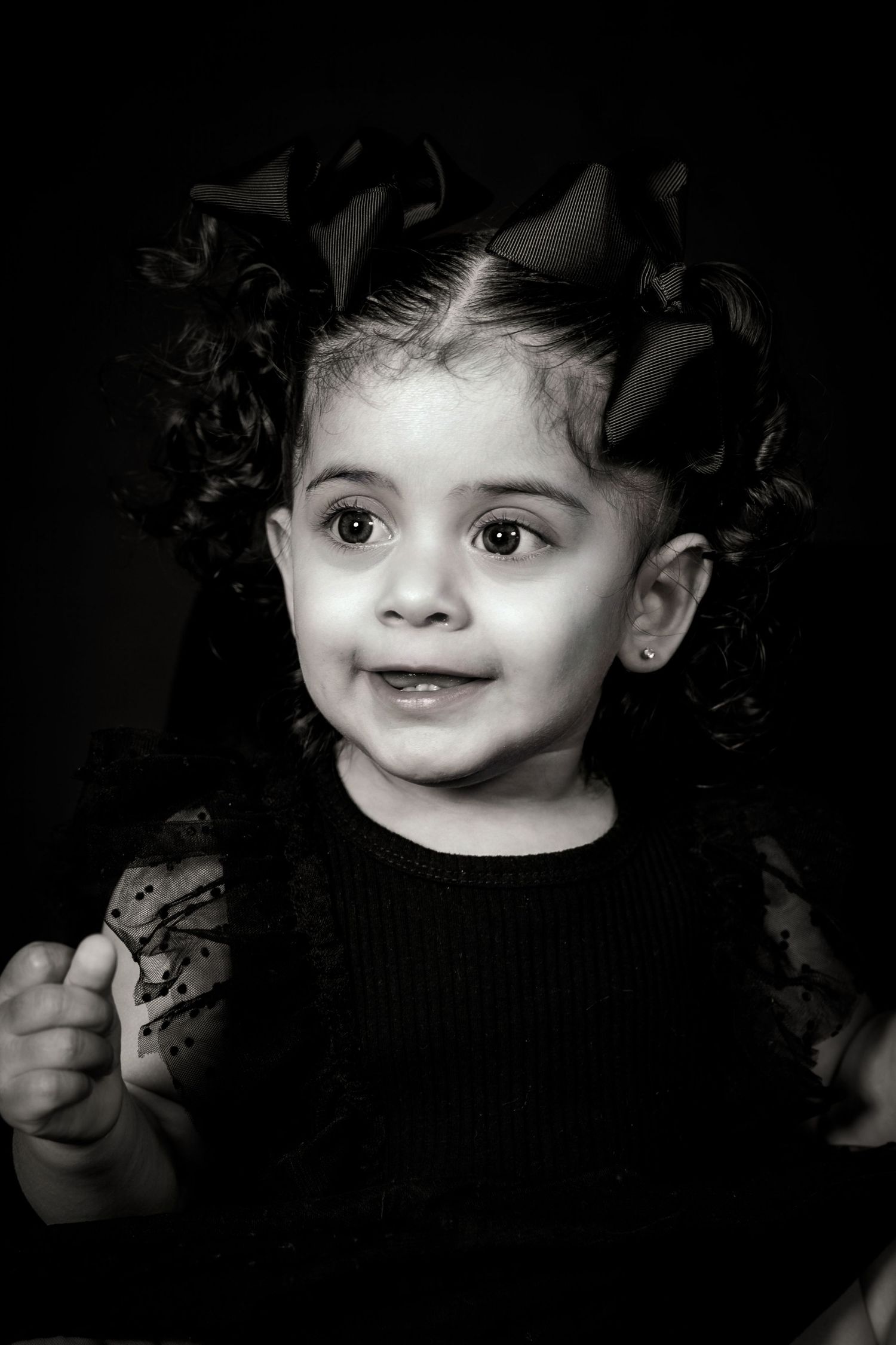 Close-up black and white portrait of toddler with curly hair and bows wearing dark clothing.