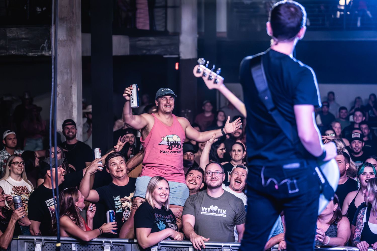 Fan in pink shirt raises fists while crowd surfing as musician performs on stage.