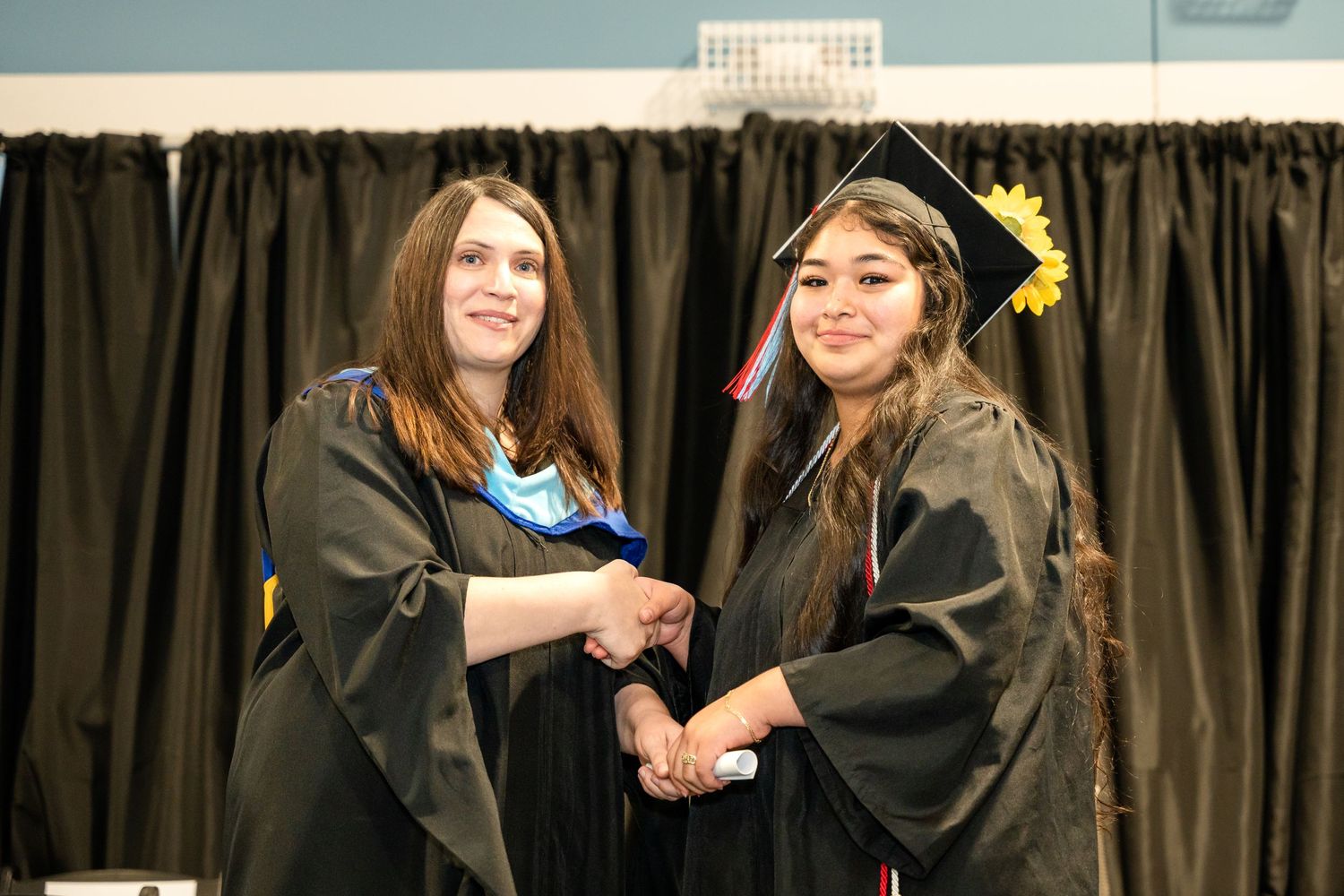 Two graduates in black caps and gowns shake hands during a graduation ceremony.