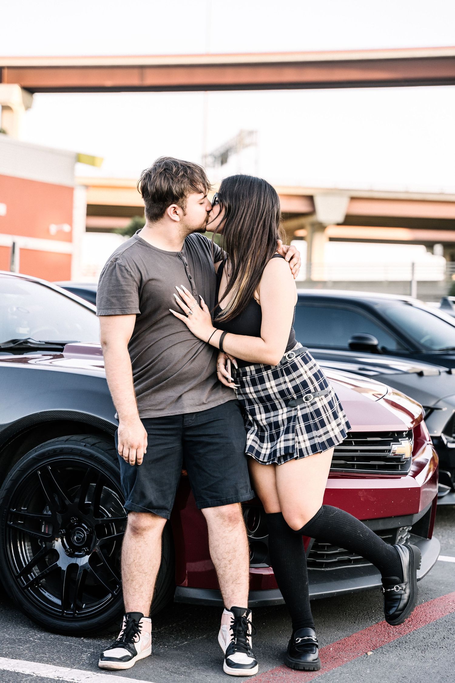 A couple in casual attire sharing a romantic moment in front of a red car in a parking lot.