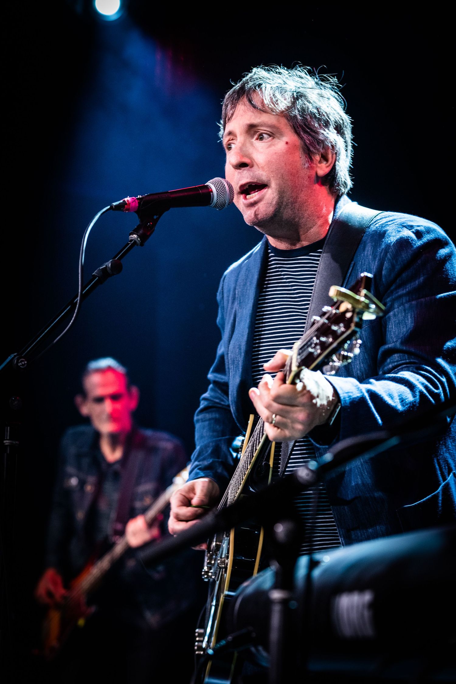 Performer in blue jacket sings passionately while strumming guitar on illuminated stage.