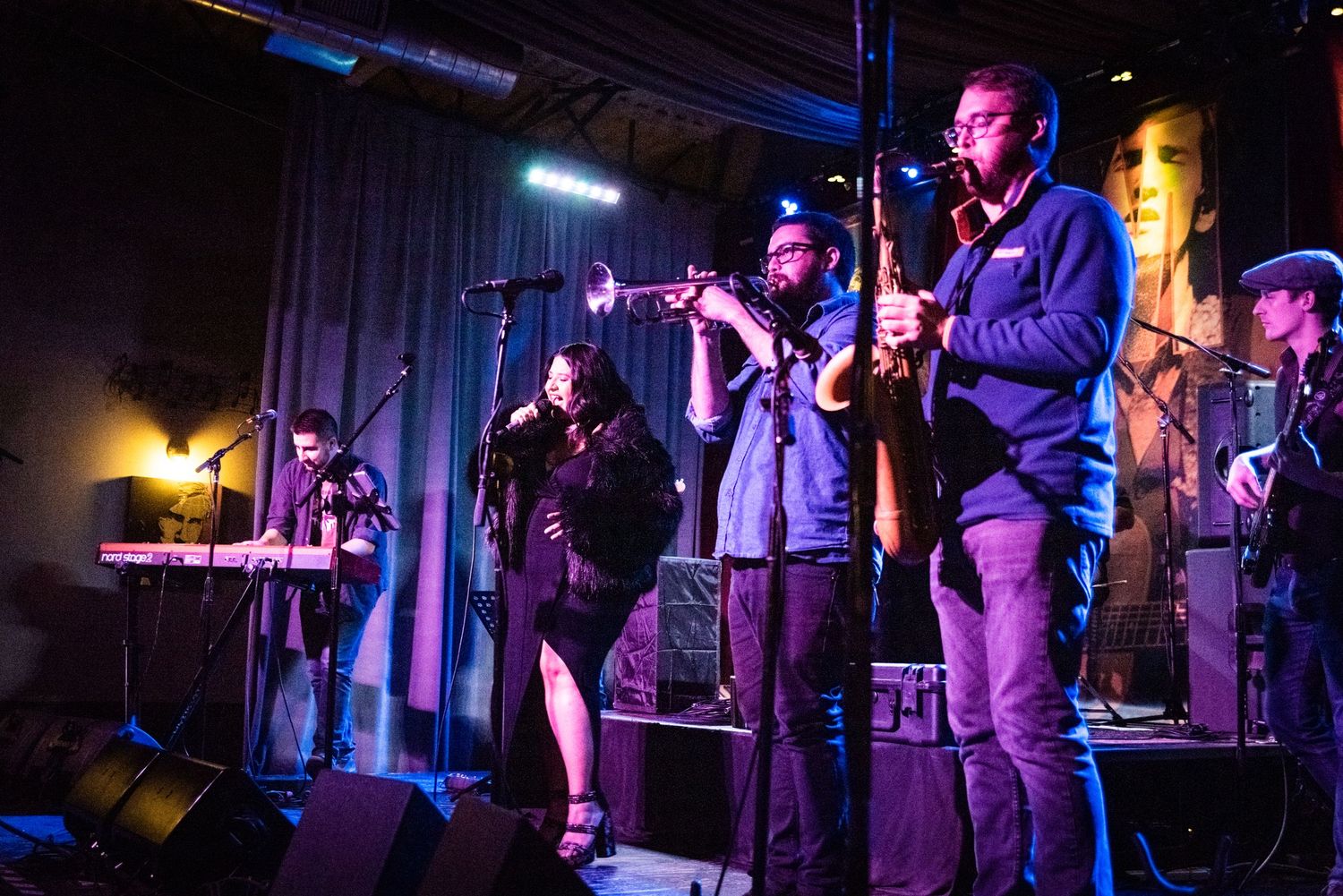 A band performs on stage with brass instruments and keyboards under blue and amber lighting.