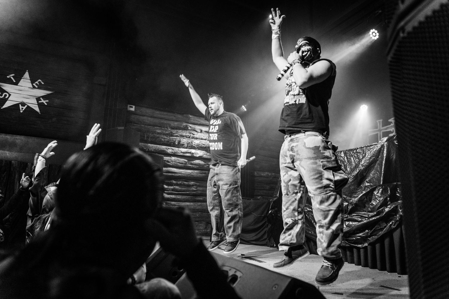 Black and white concert photo showing performers on stage with audience silhouettes.