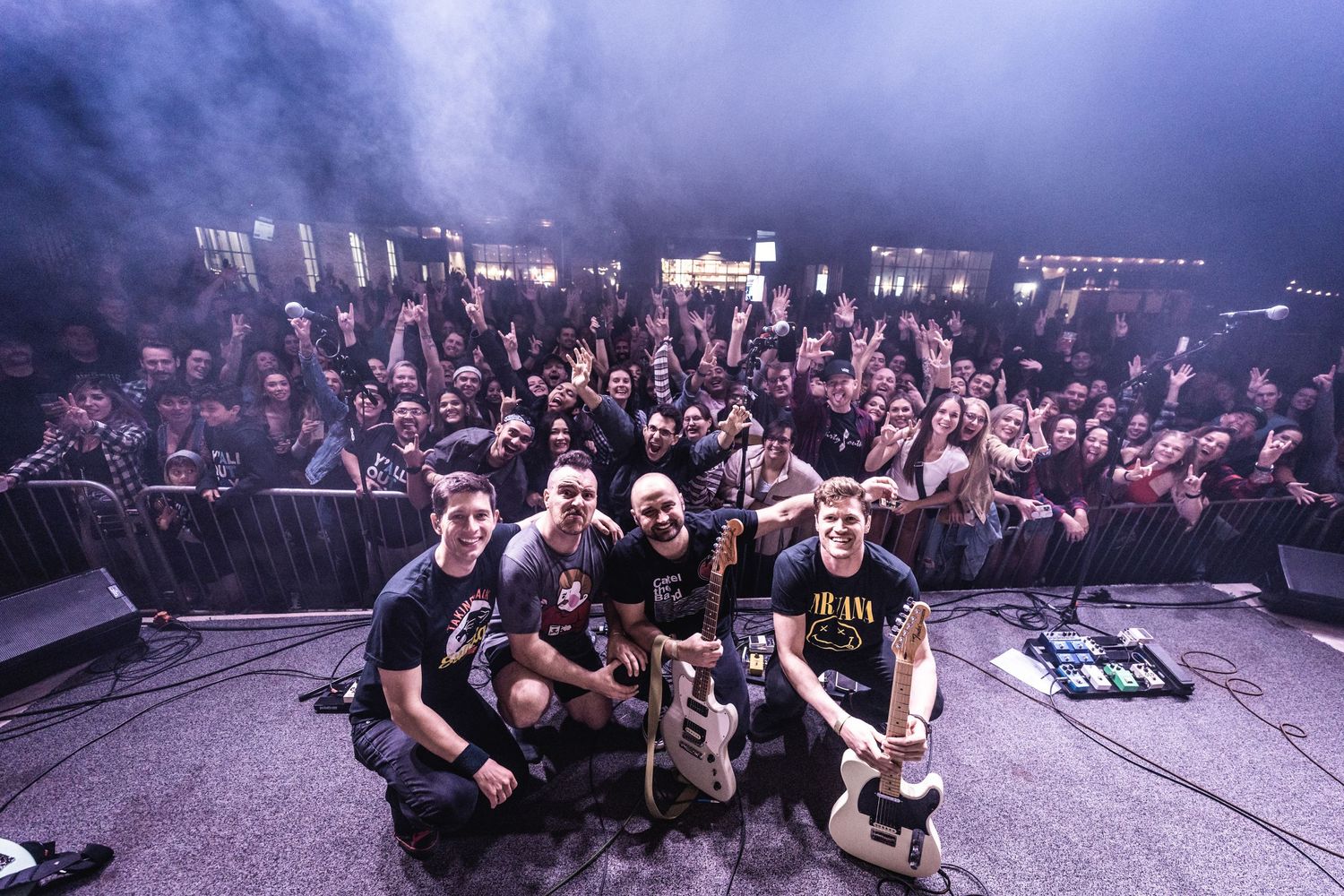 Band and crew members pose for group photo on stage with crowd visible behind.