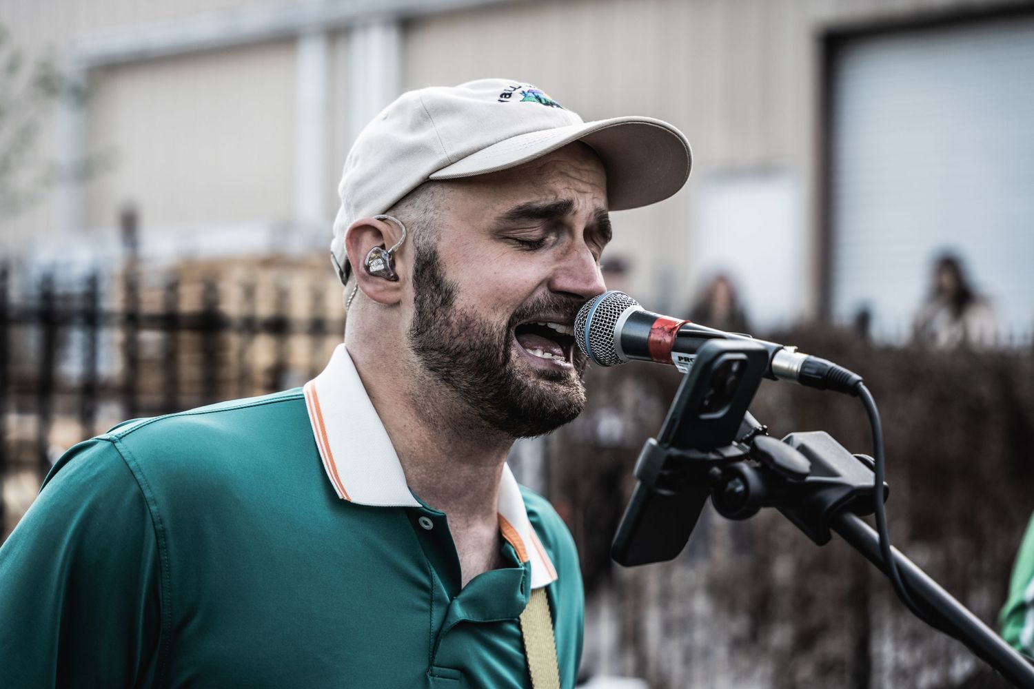 Singer in green polo and cap performs with microphone during outdoor event.