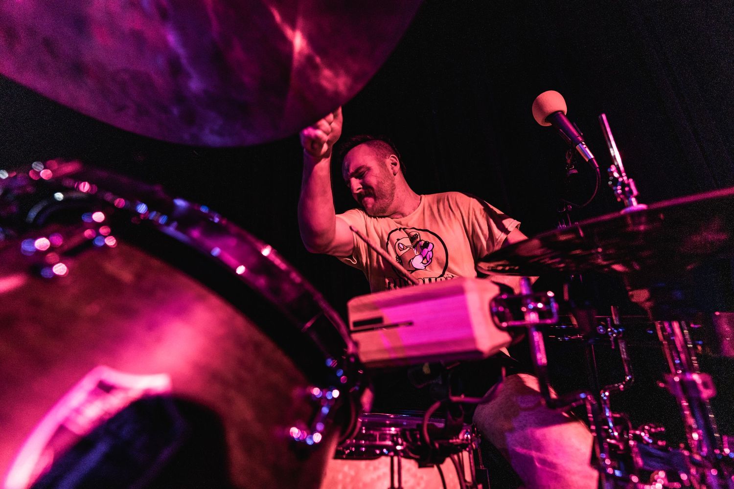 Energetic drummer performs on stage bathed in dramatic pink and purple lighting.