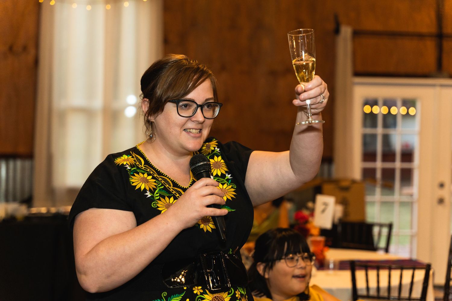 Guest raises champagne glass for wedding toast wearing floral dress at reception.
