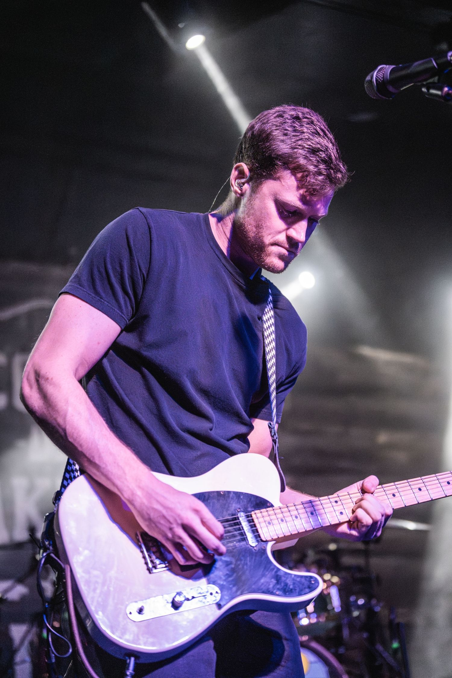 Guitarist in navy shirt performs under bright spotlights on dark stage.