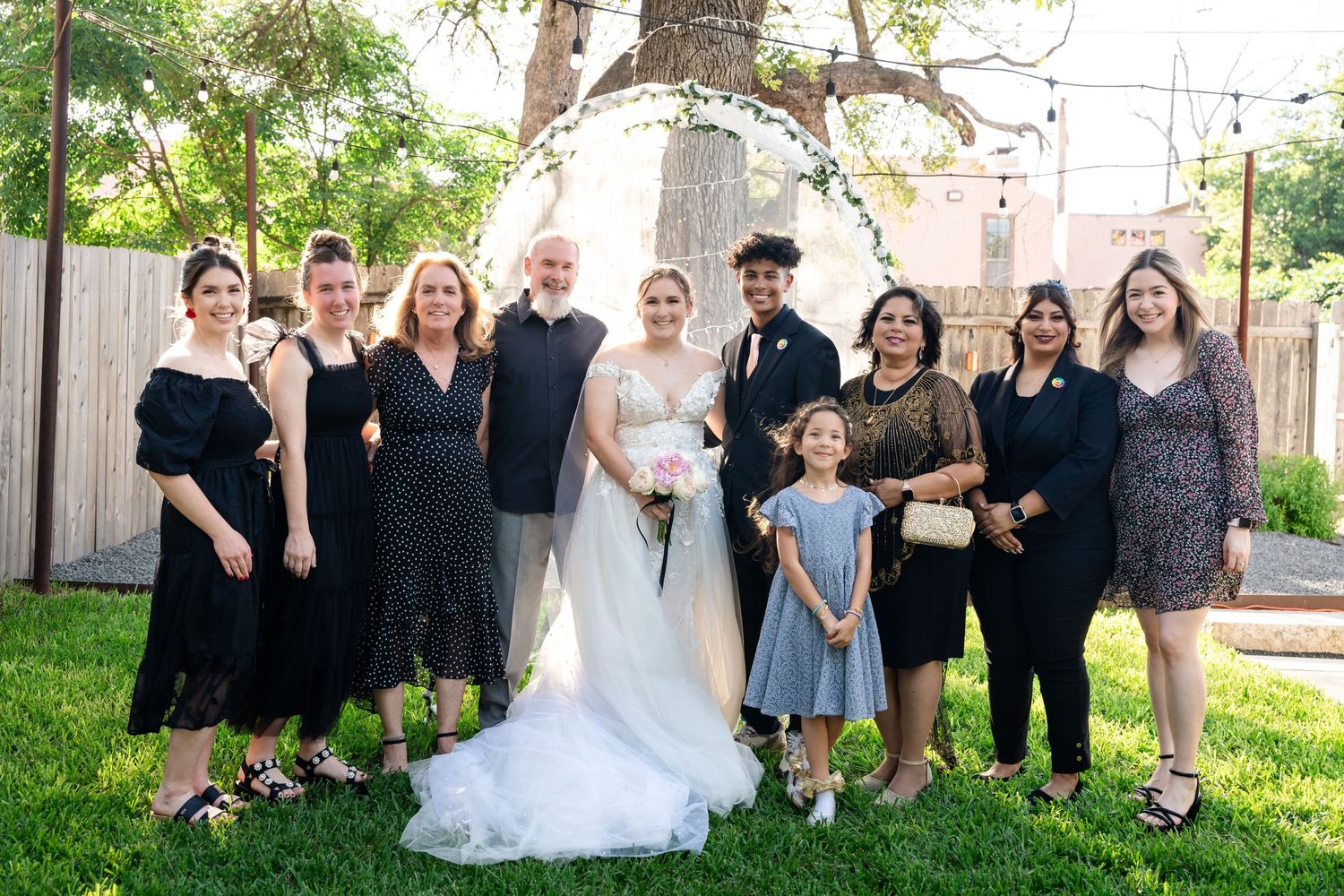 Wedding party poses together outdoors in mix of formal attire on sunny day with wooden fence backdrop.