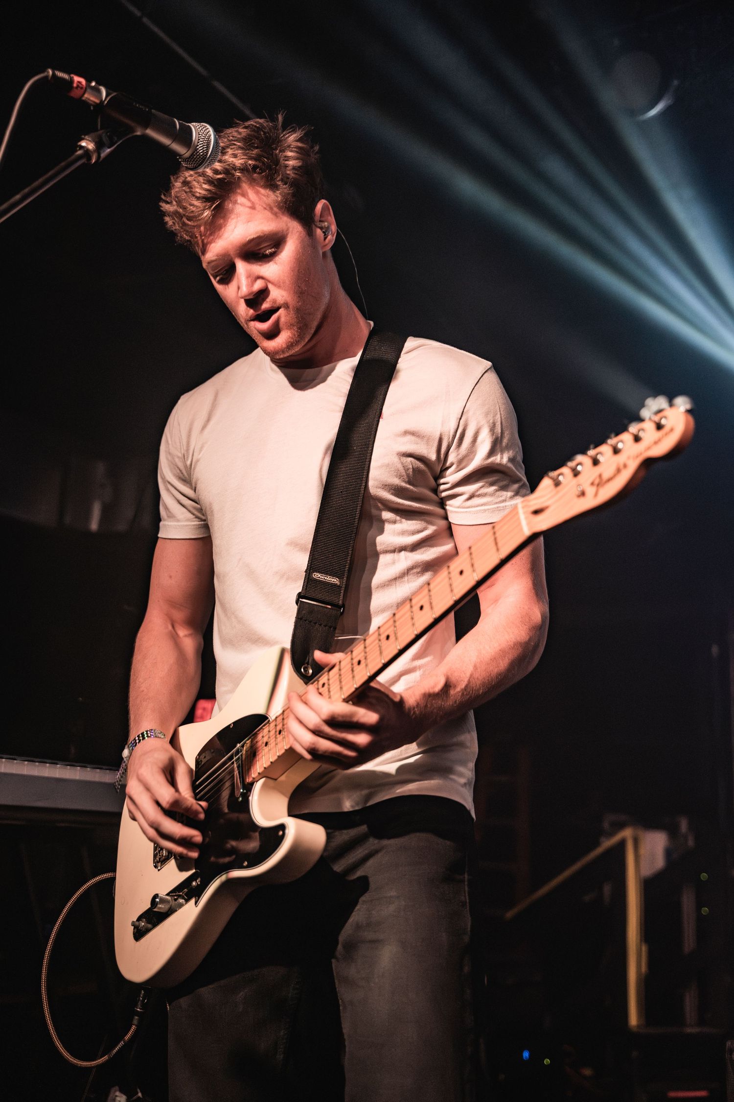 A guitarist in a white t-shirt performs on stage with dramatic blue lighting and beams in the background.