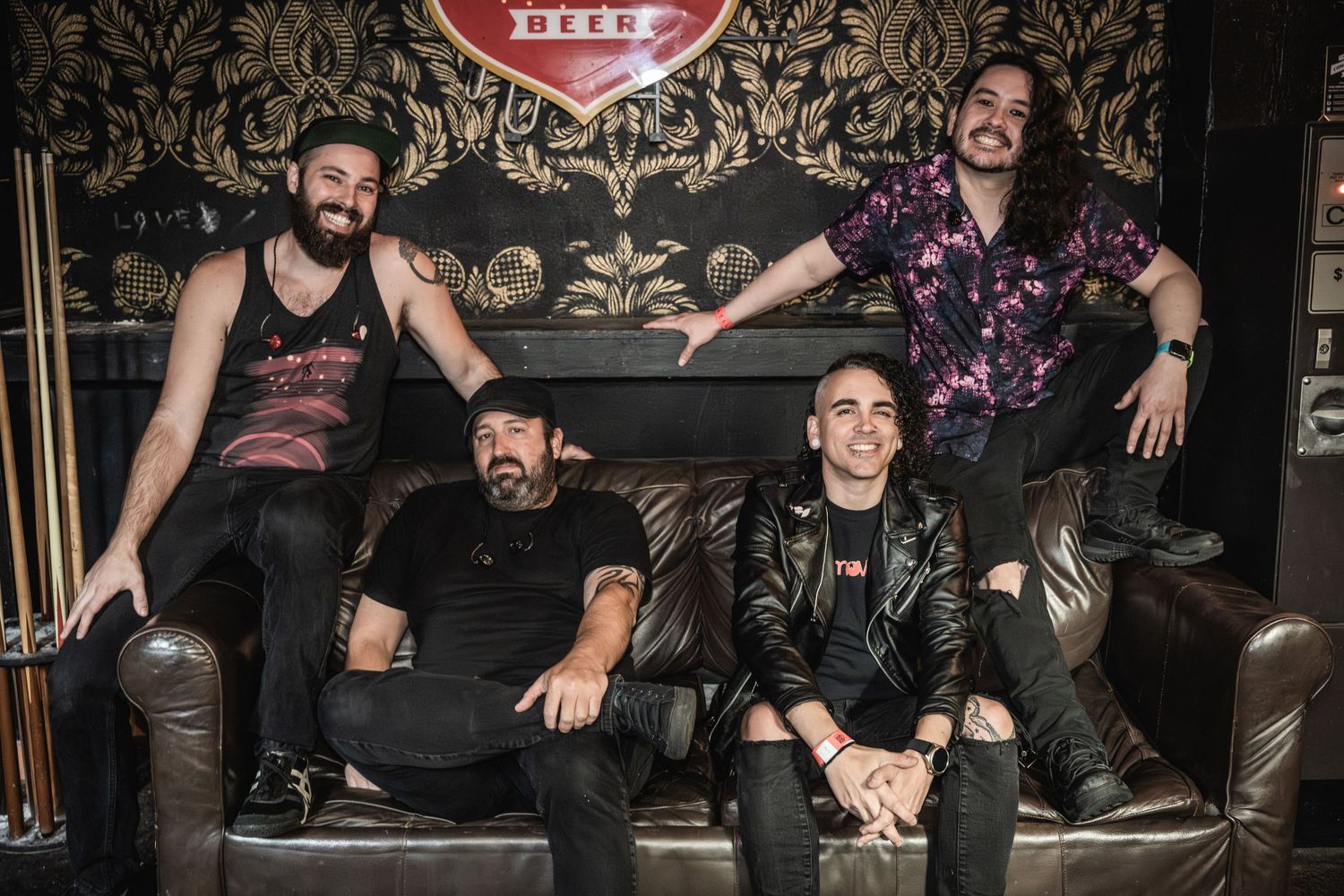Rock band members posing on leather couch against ornate wallpaper in dark venue setting.