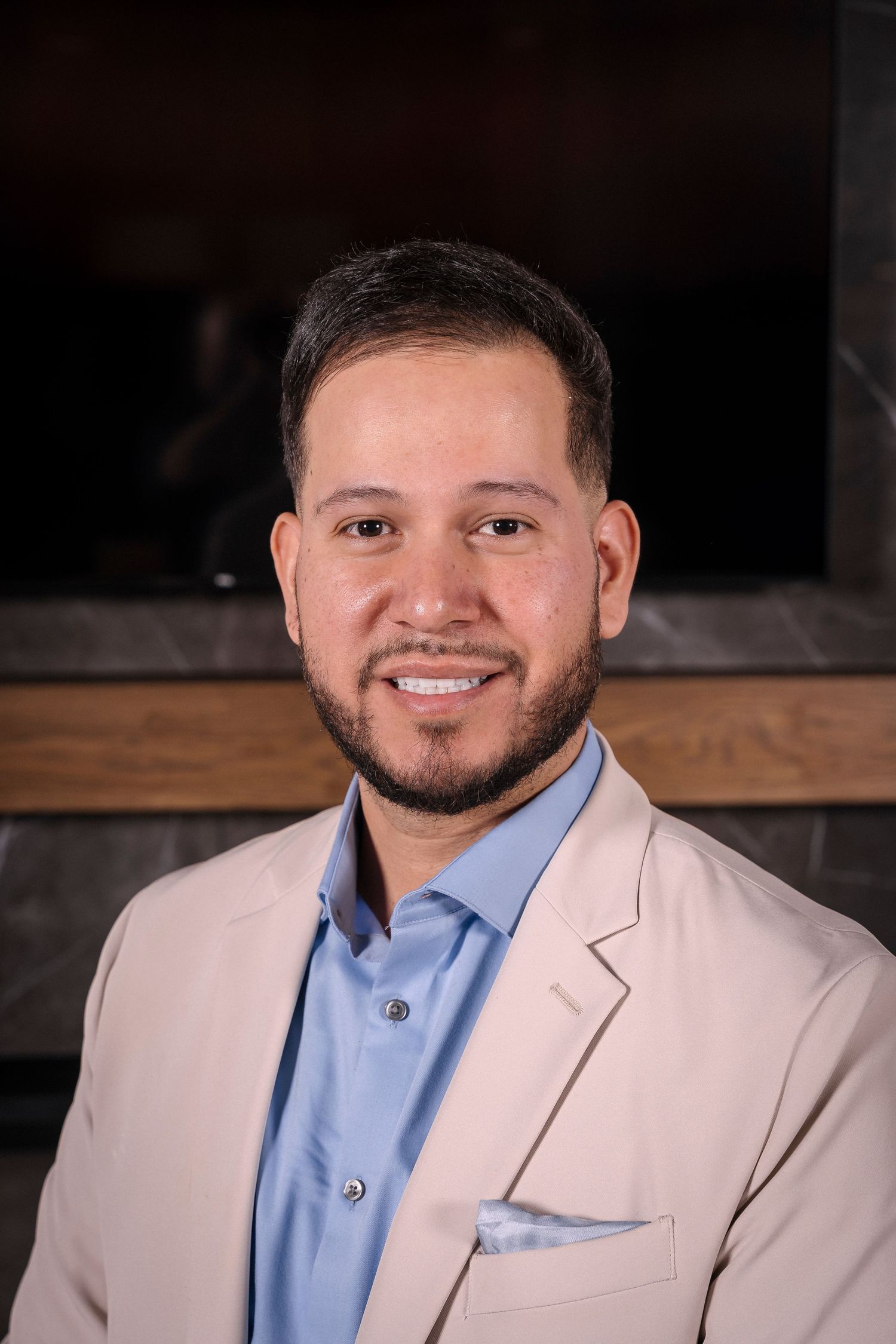 Professional headshot portrait of someone wearing a light beige blazer and blue dress shirt against dark background.