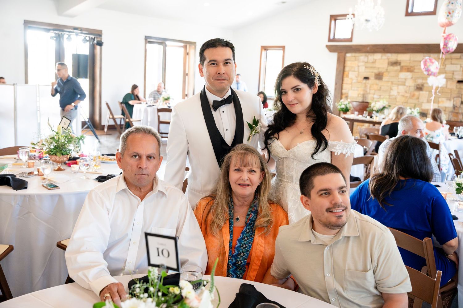 A group of guests seated at a white-clothed table during a wedding reception in a rustic venue with stone walls.