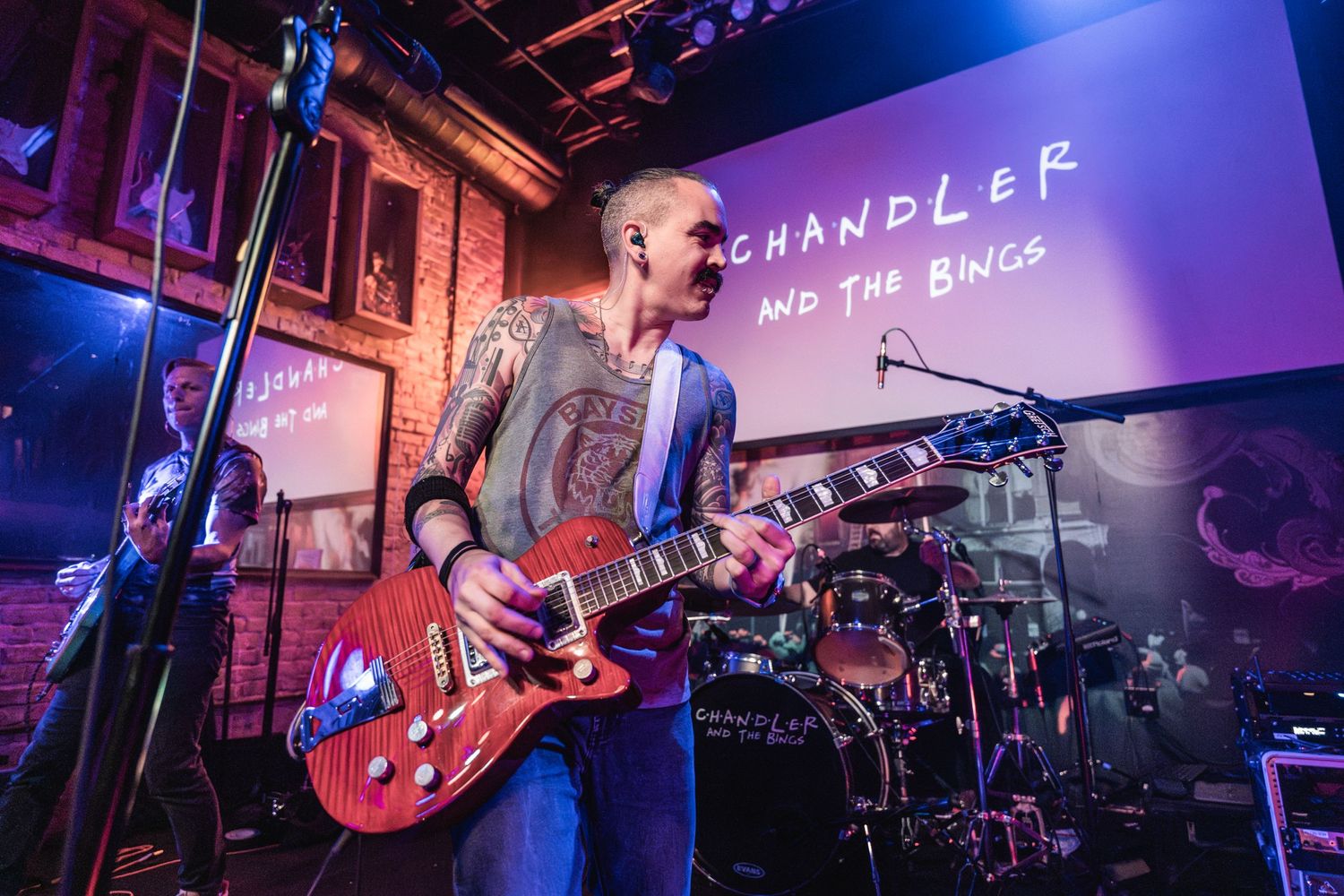 A musician performs on stage with a red guitar beneath a projection screen displaying 'Chandler'.