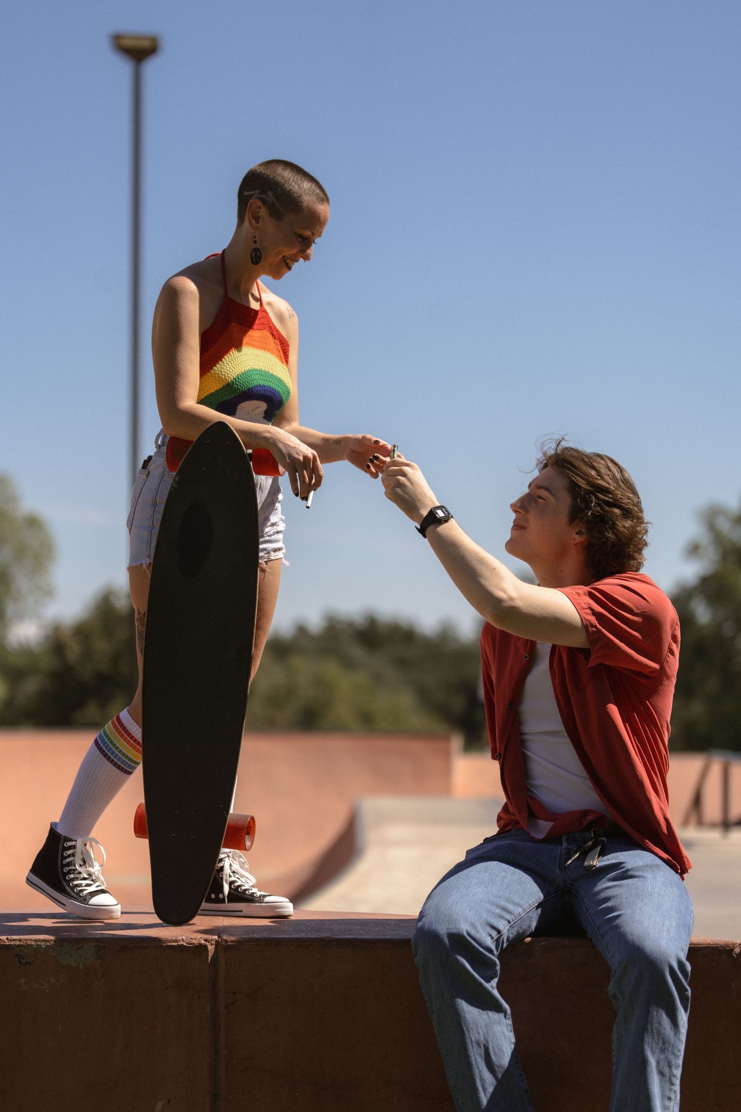 Two people share a moment at a skate park, one standing on a ledge with a skateboard.