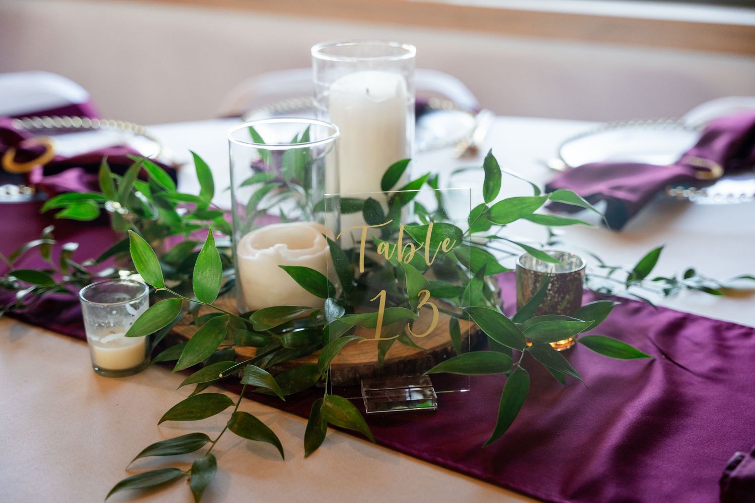 Table centerpiece with white candles and greenery on burgundy table linens.