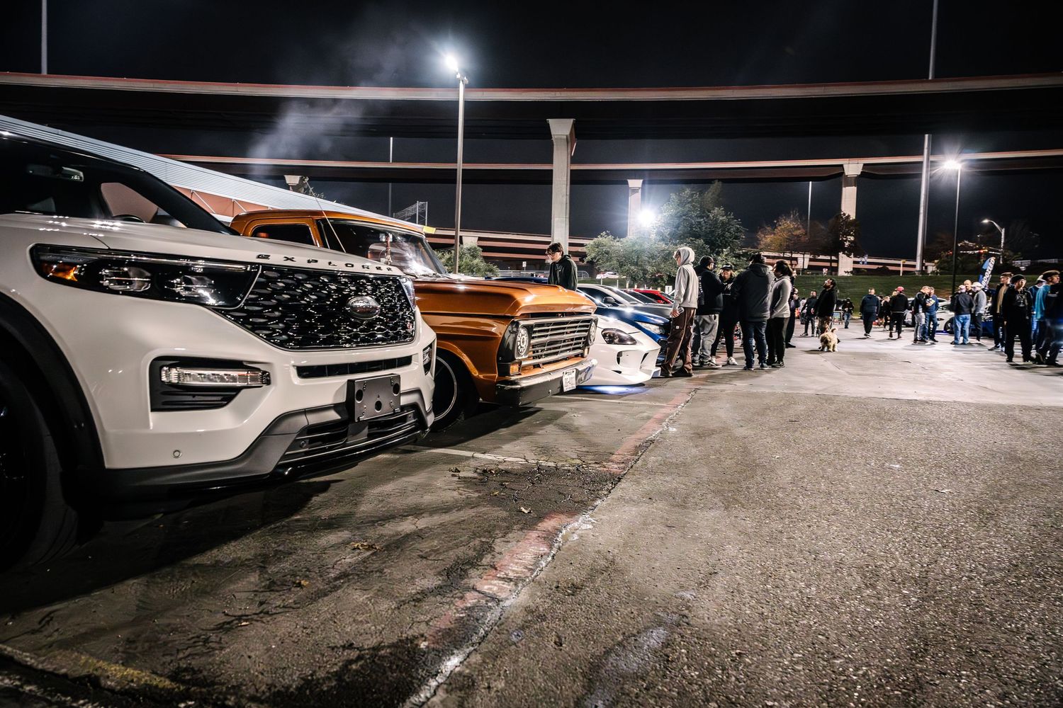 Line of modified vehicles at a nighttime car meet with crowd gathered in the background.