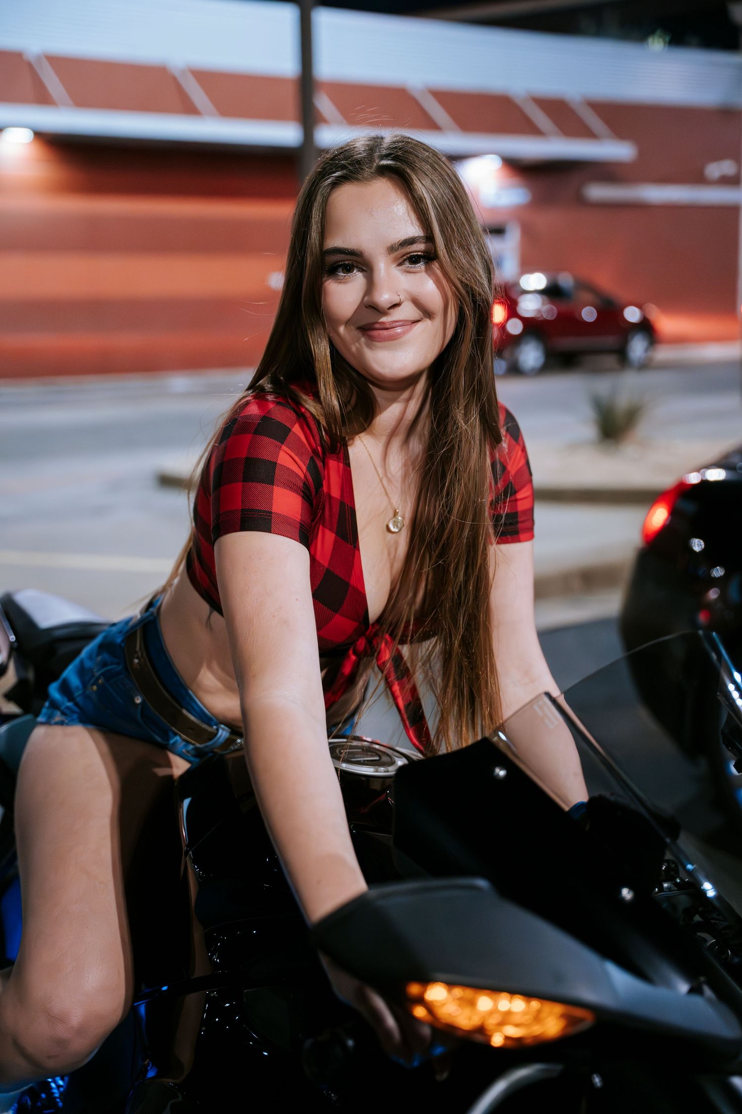 Model in red plaid top poses on black motorcycle at outdoor car event.