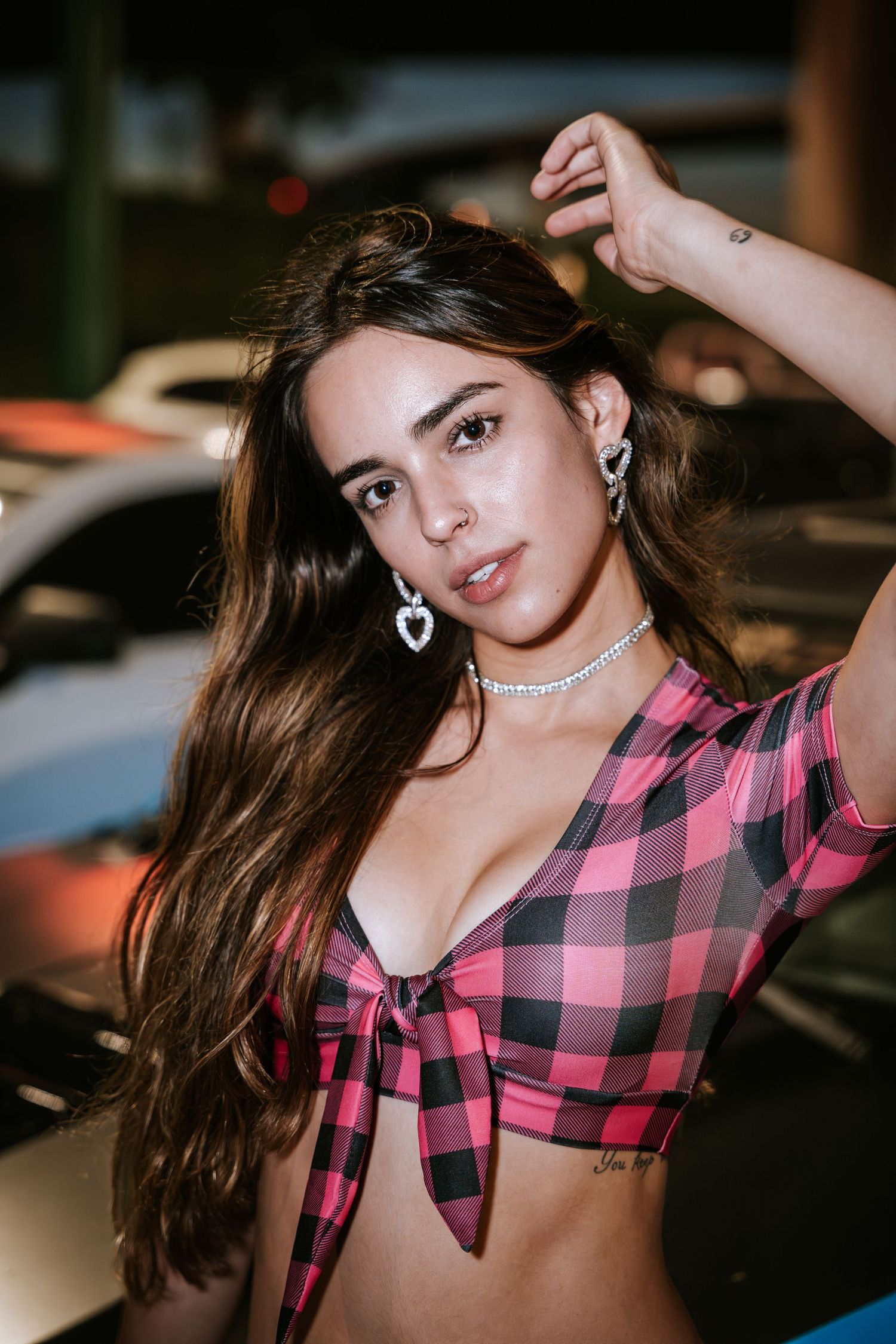 Model in pink plaid crop top and heart earrings poses against cars in parking lot at night.