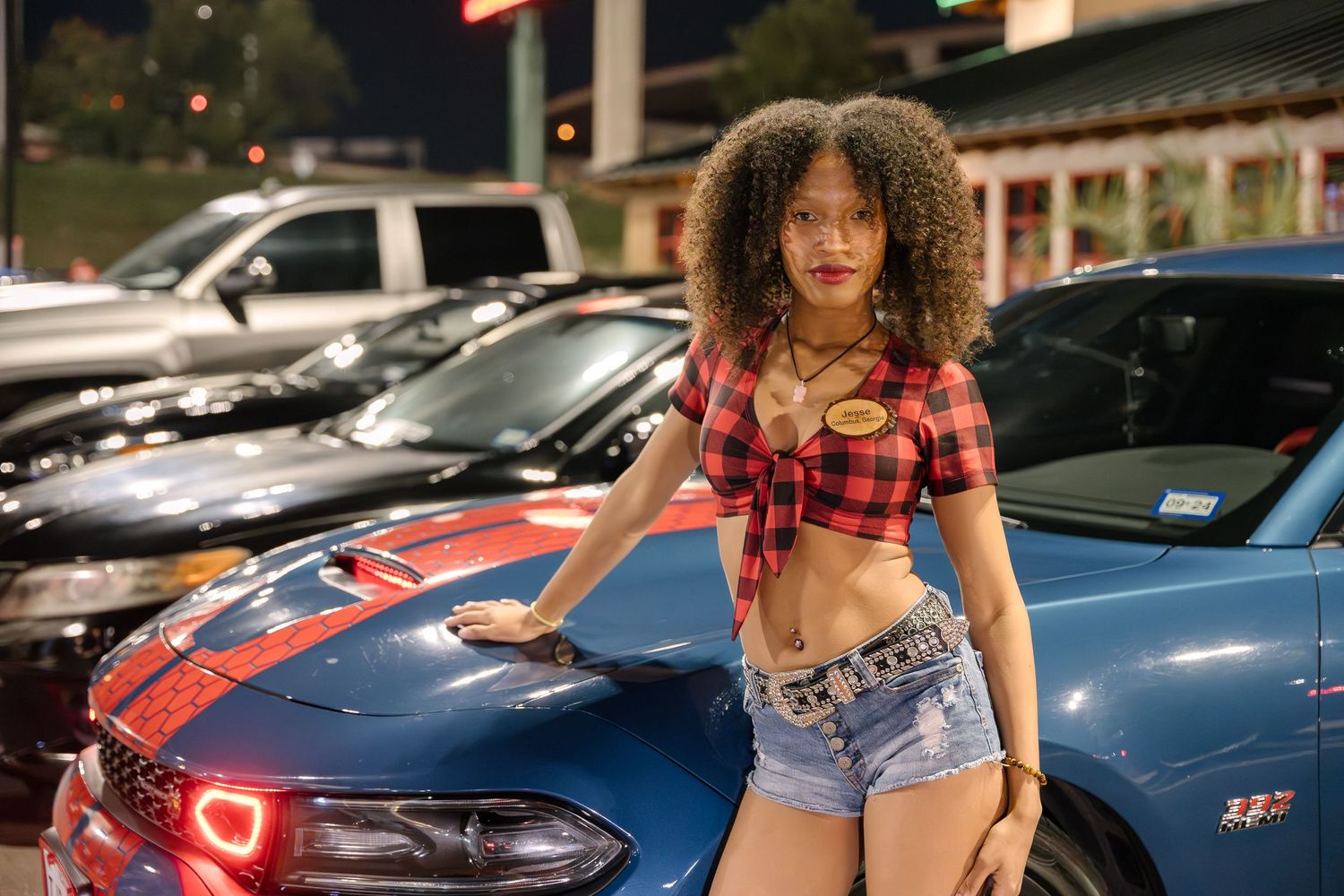 Woman standing beside blue sports car at outdoor nighttime car show.
