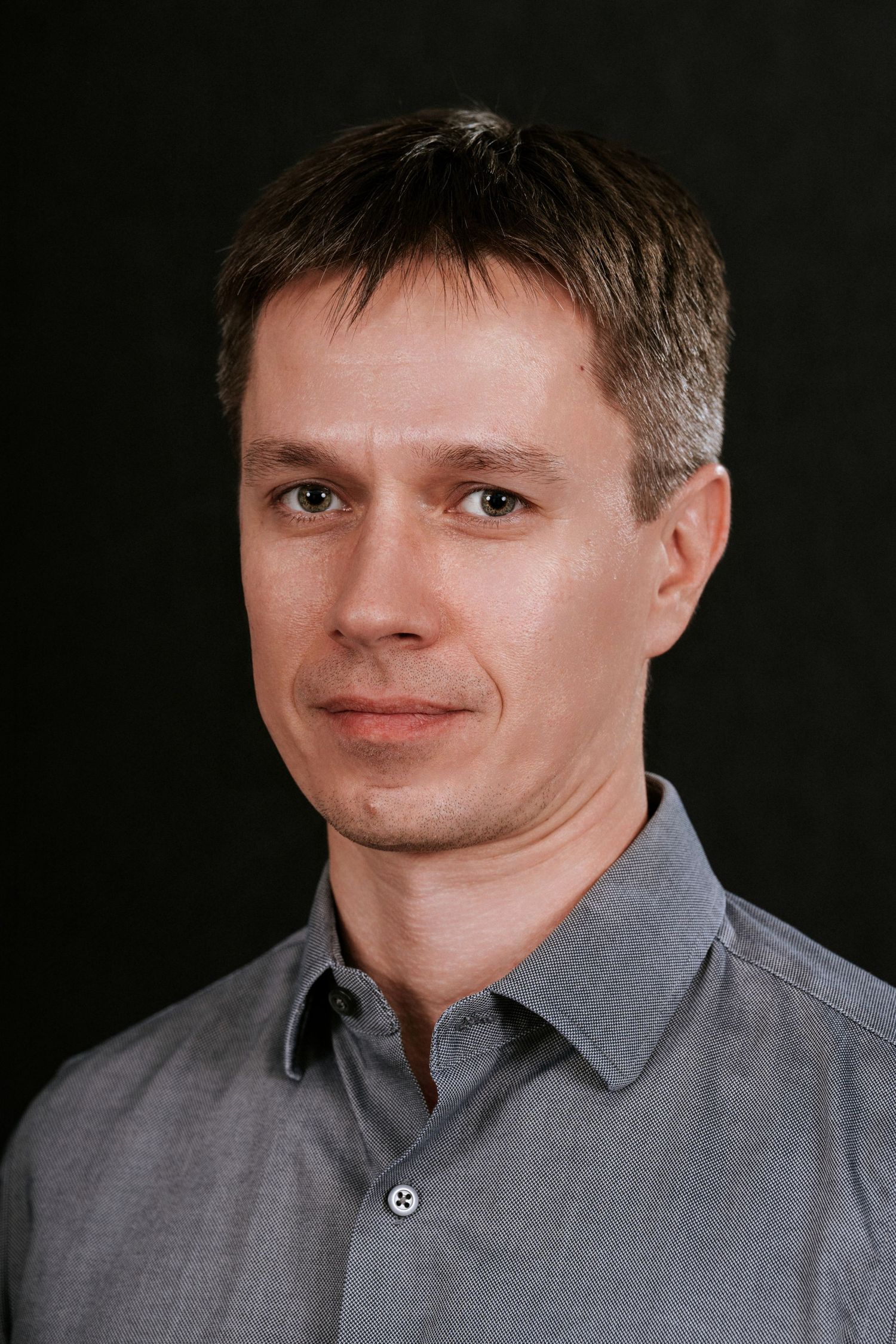 Studio headshot of person wearing grey collared shirt against dark background.