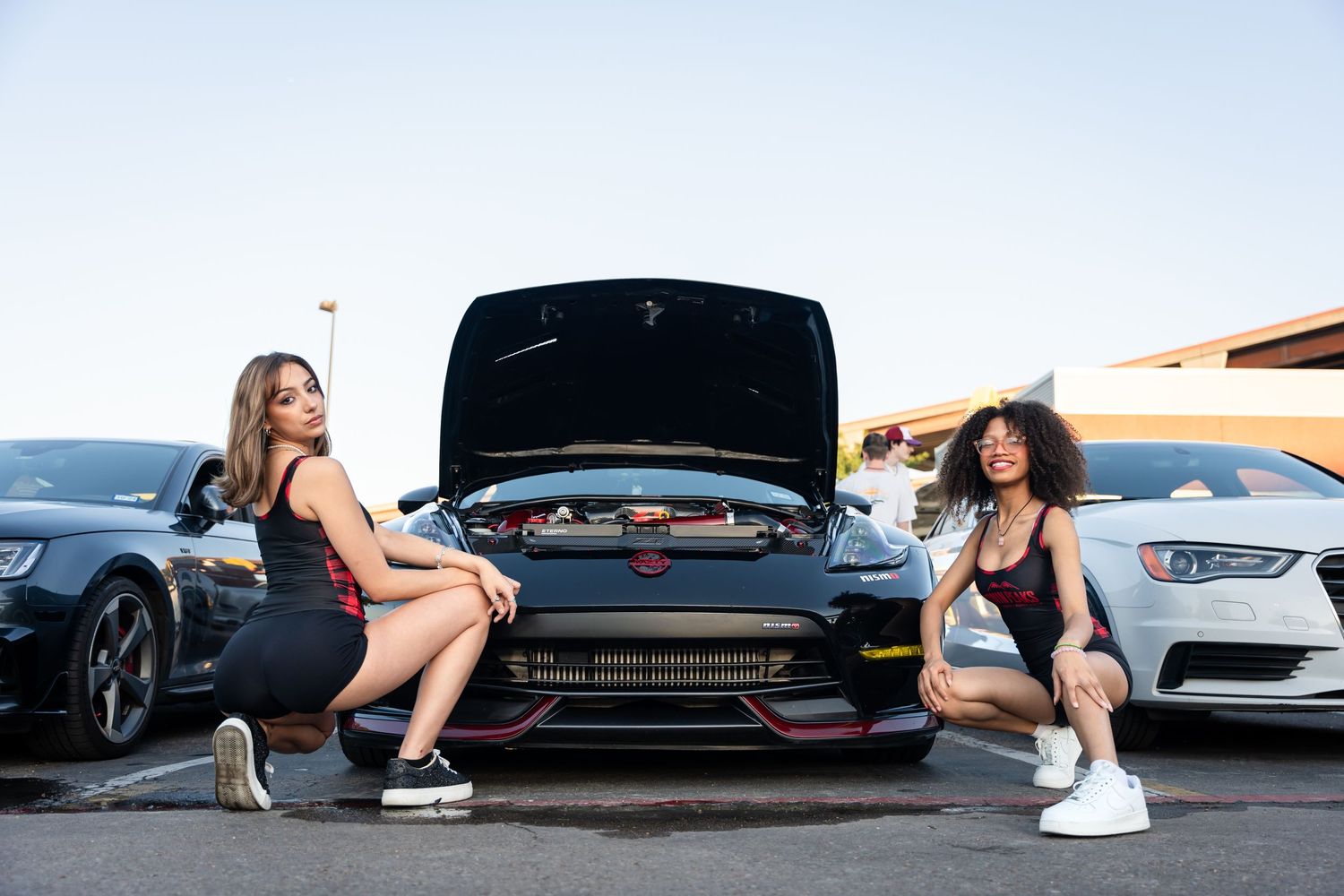 Two models in matching outfits pose near car with open hood at auto meet.