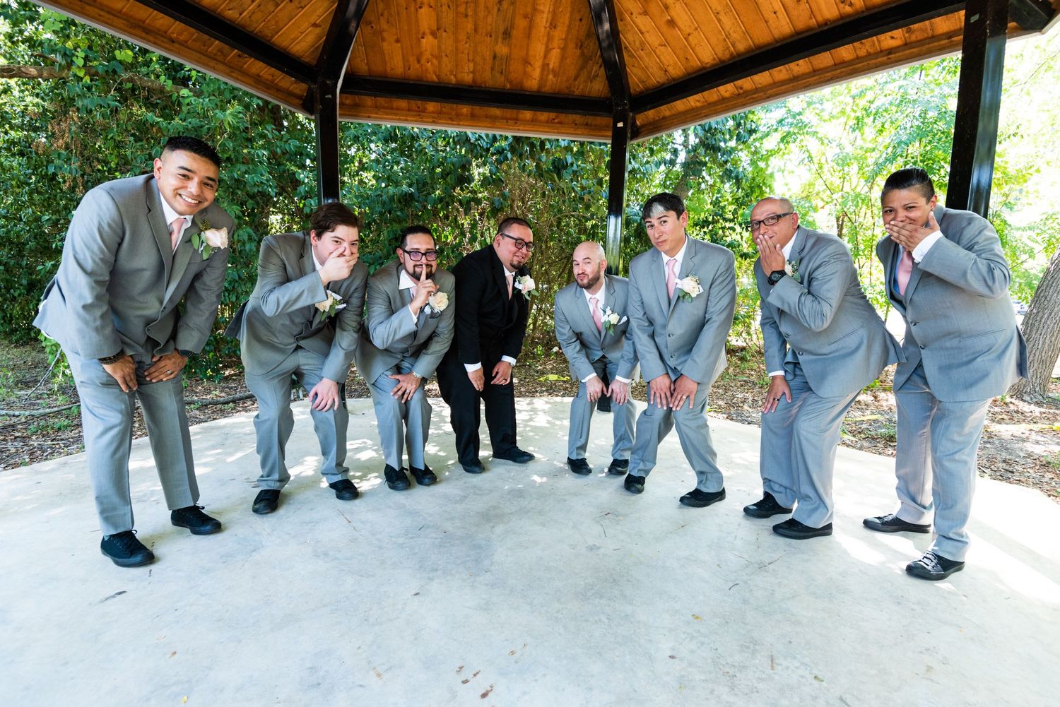 Groomsmen in grey suits pose playfully under wooden gazebo showing off their watches.