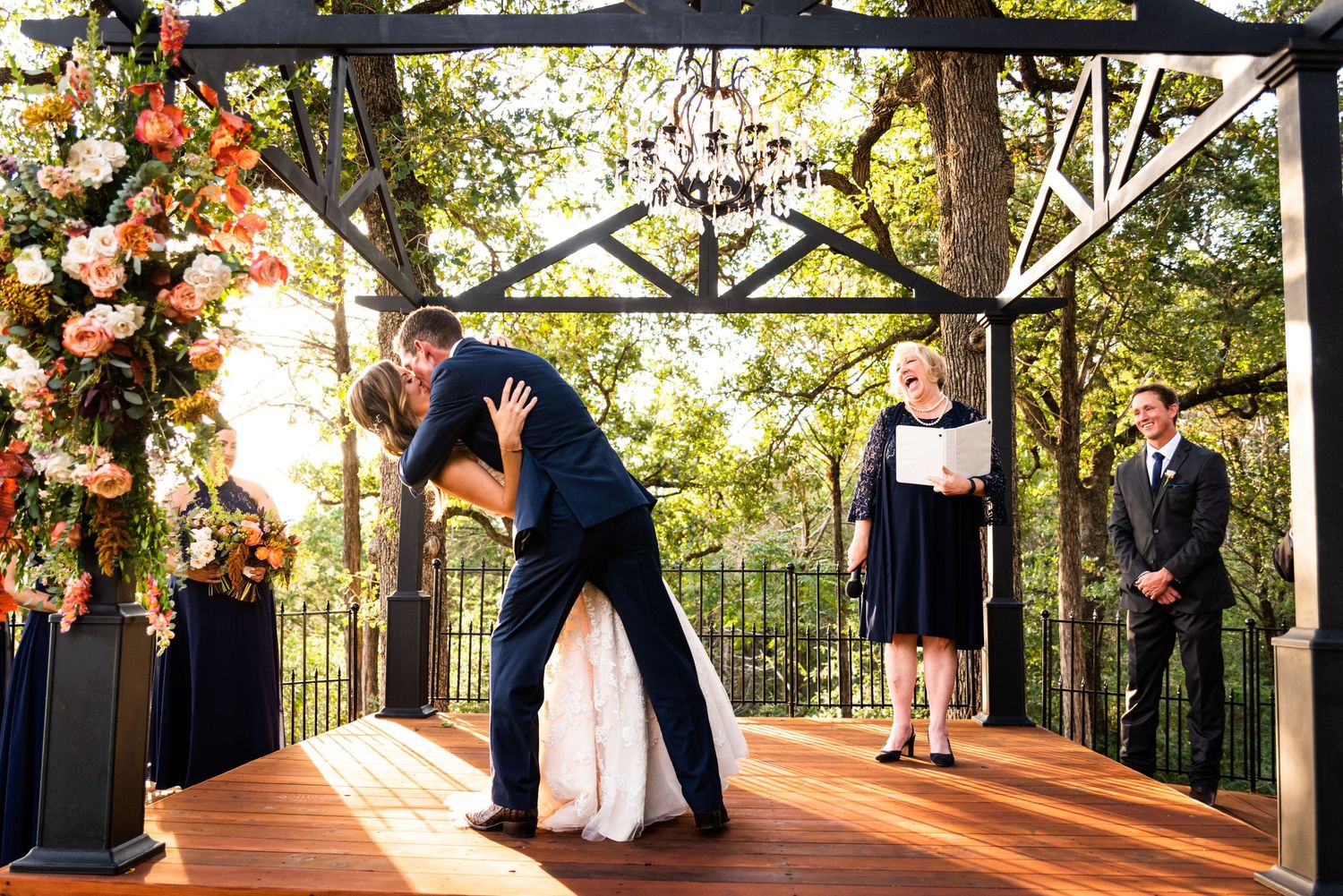 A dramatic wedding ceremony kiss on a wooden deck decorated with floral arrangements and chandeliers.