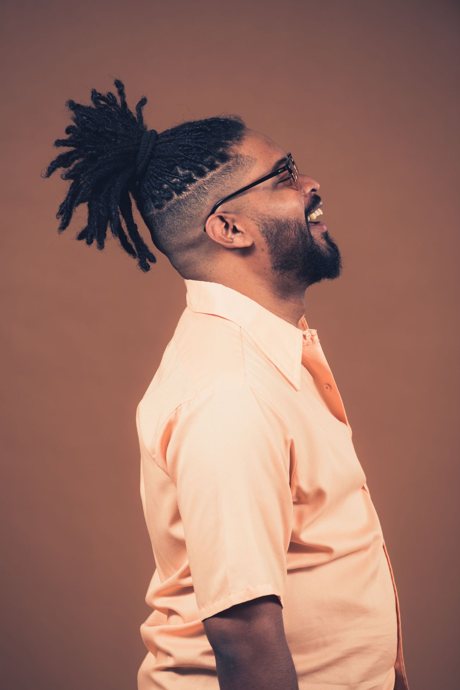 Profile view of person with dreadlocks in peach shirt against brown background.