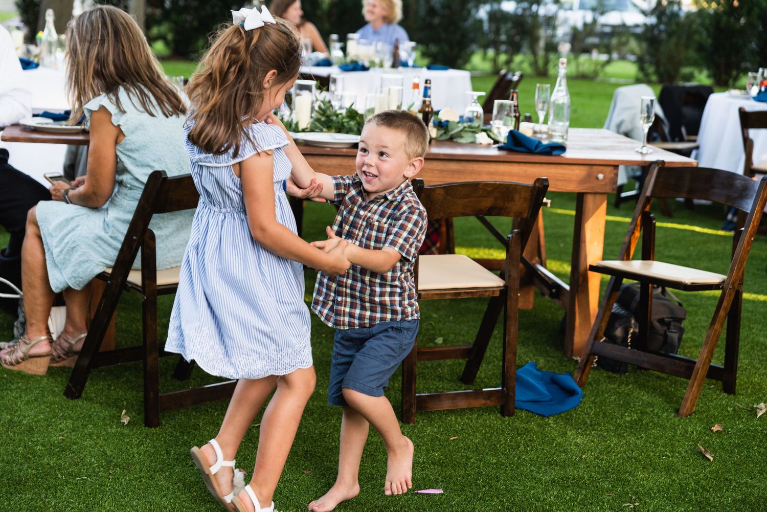 Two children dancing together at an outdoor wedding reception with wooden tables in background.