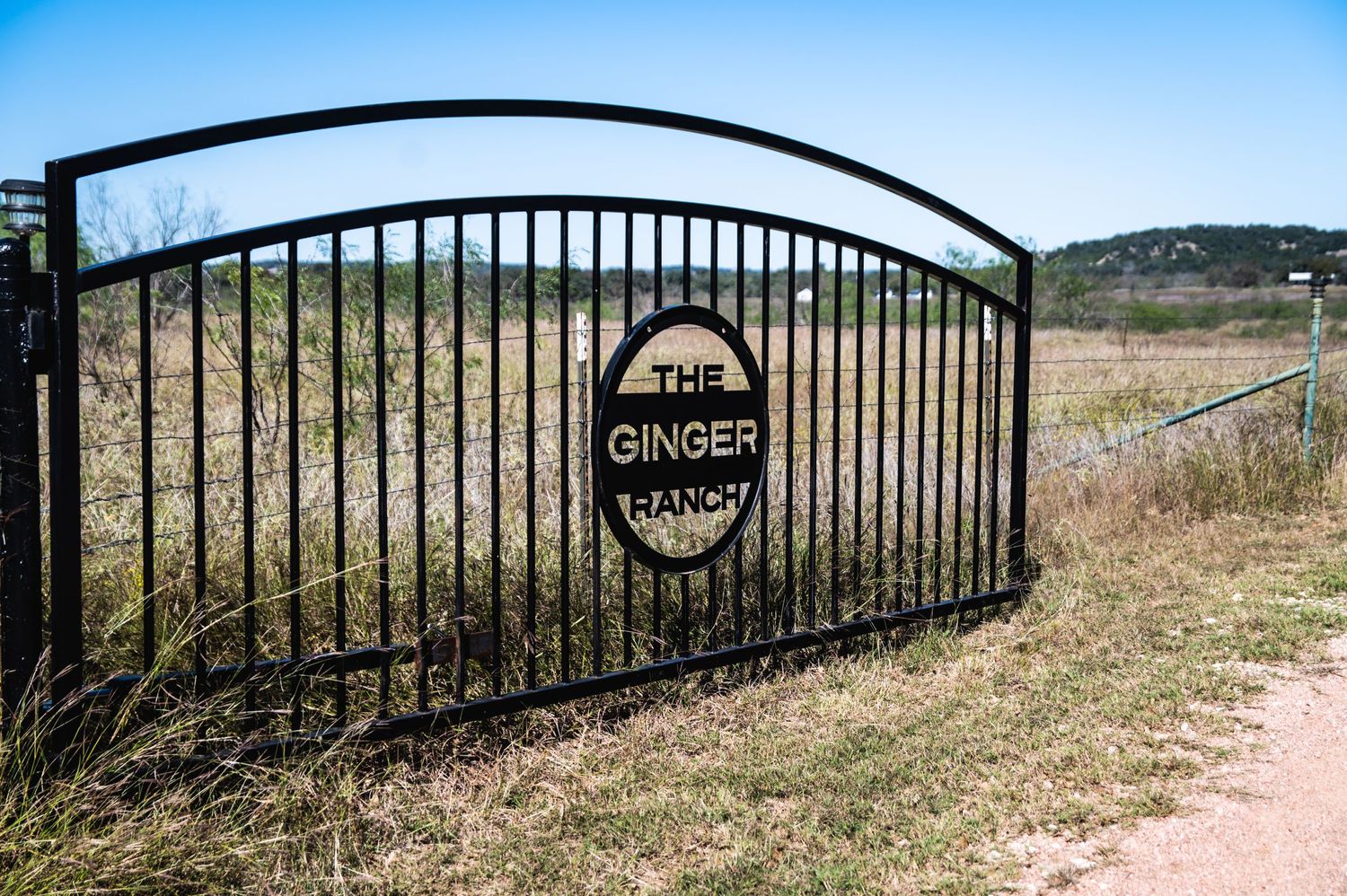 Black metal entrance gate with Ginger Ranch signage against a blue sky and rural landscape.