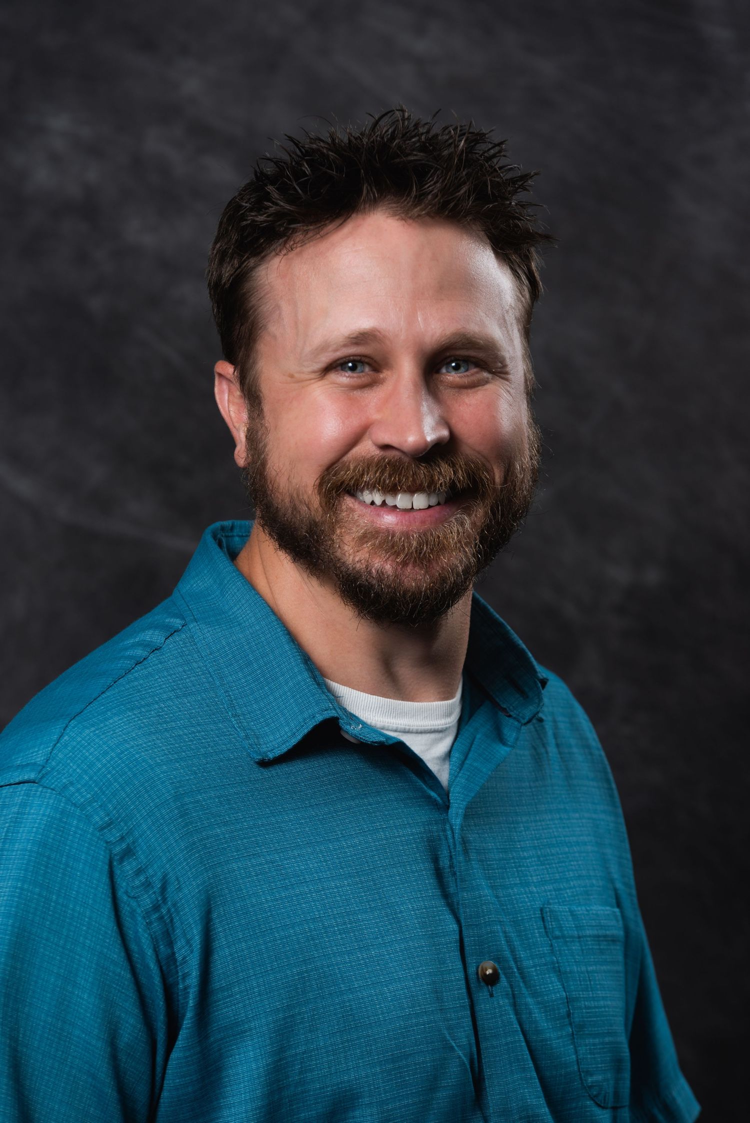 Studio portrait featuring a bearded person in a teal button-up shirt against a dark backdrop.
