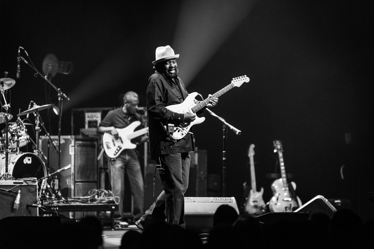 A black and white concert photo showing a musician playing electric guitar on stage with equipment in background.