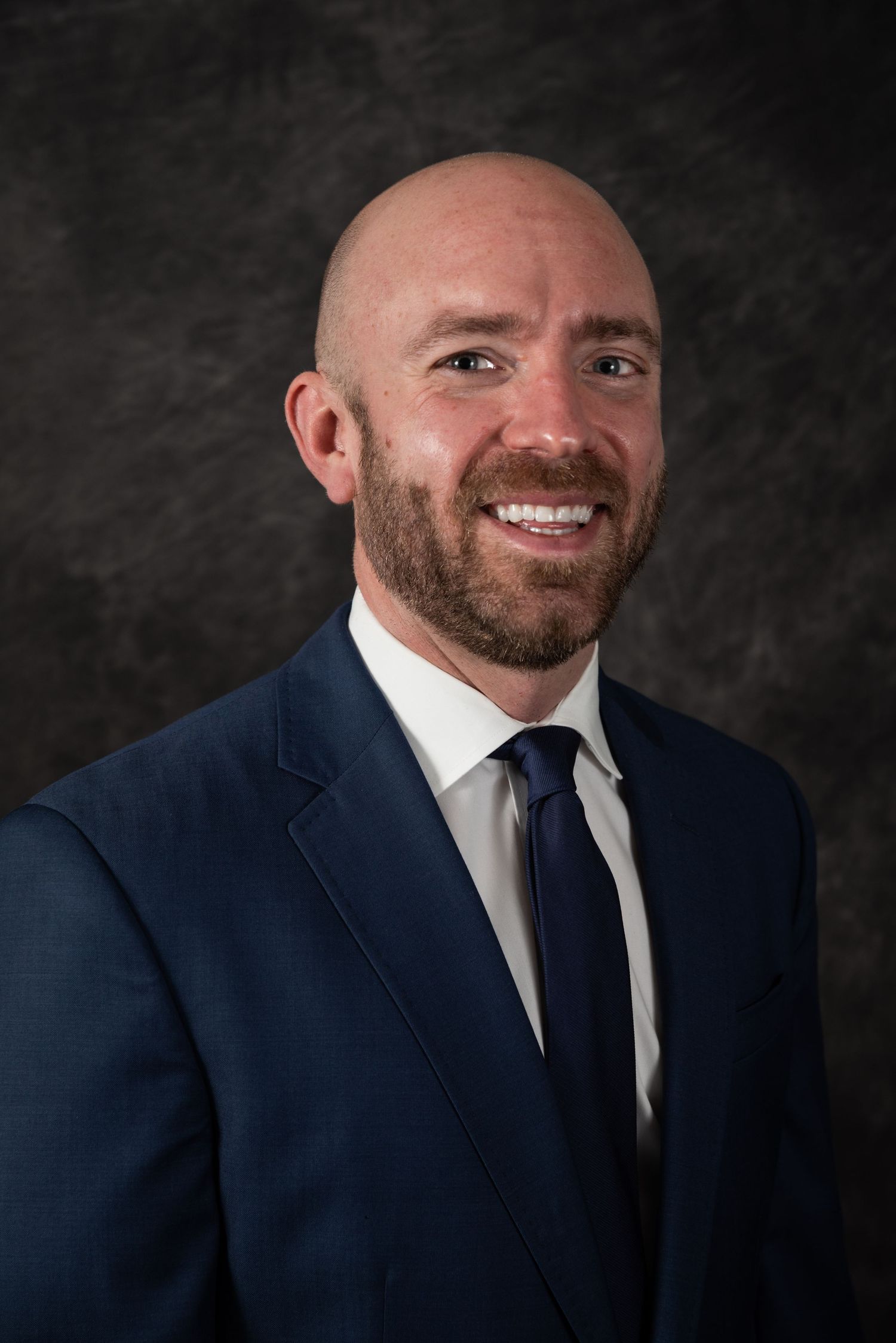 Professional portrait in navy suit and tie against textured dark background.