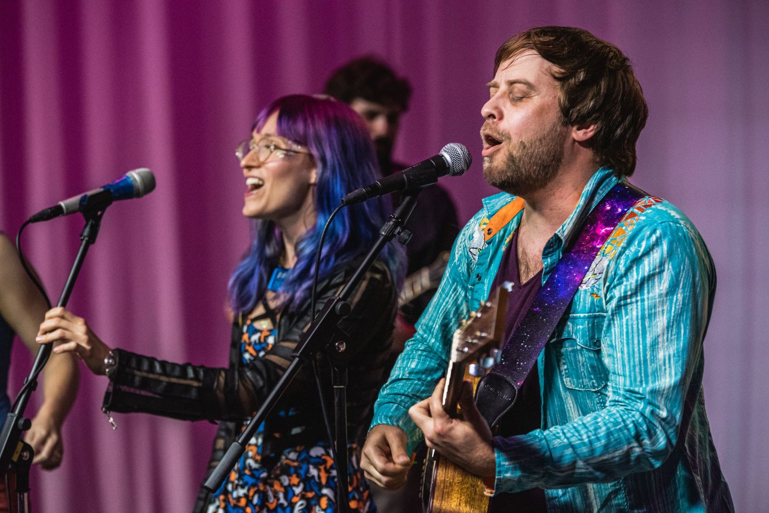 Musicians with guitars performing on stage with pink backdrop.