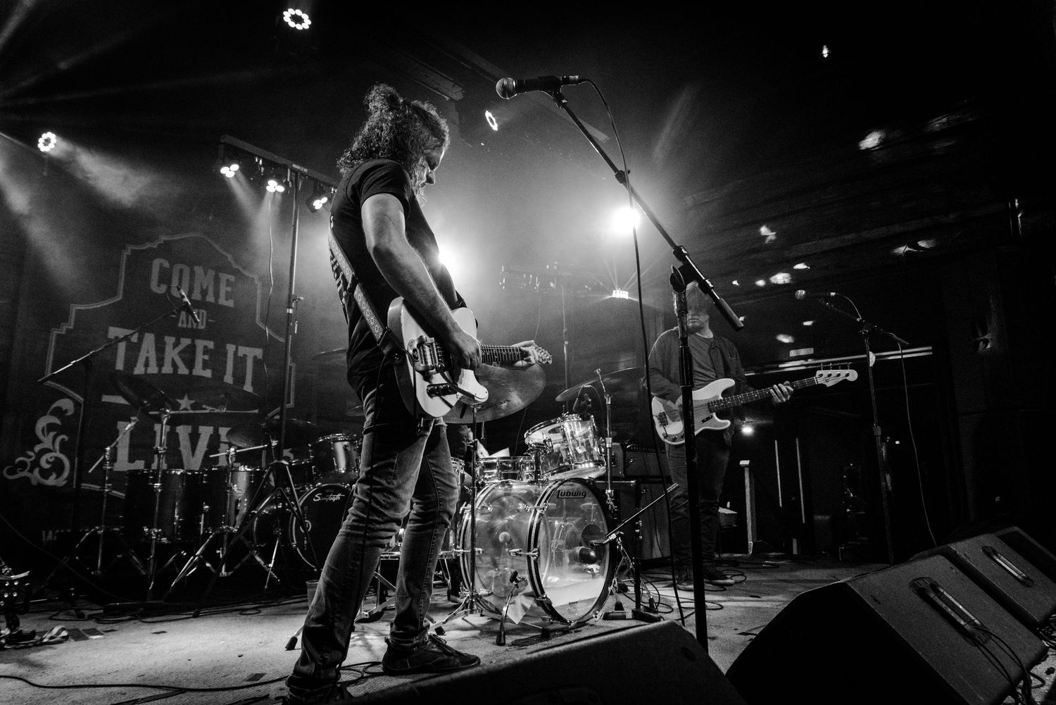 Black and white photo of a musician with guitar performing on stage with dramatic lighting and drum kit in background.