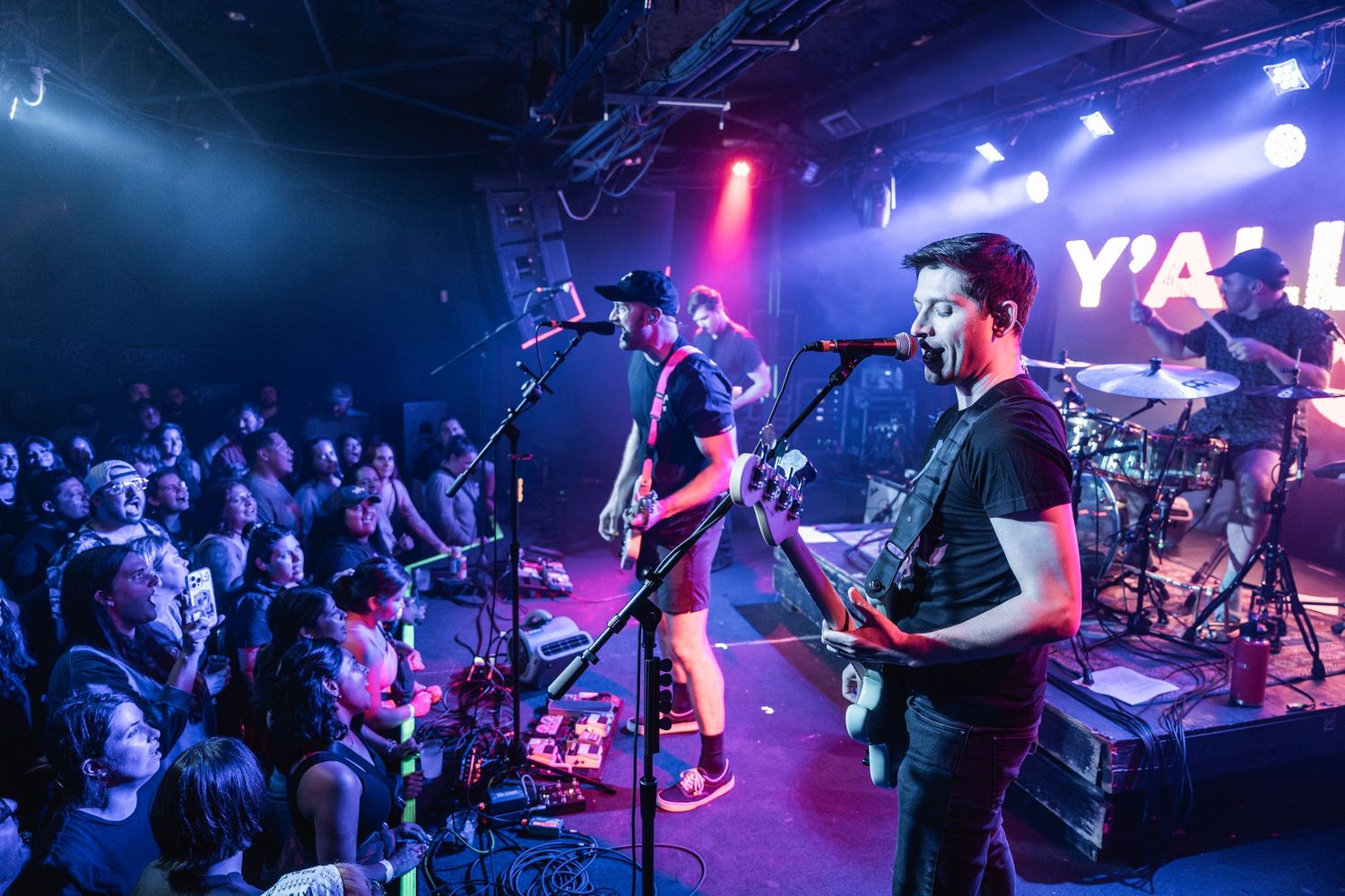 Musicians perform on stage with colorful blue and pink lighting effects at a live concert venue.