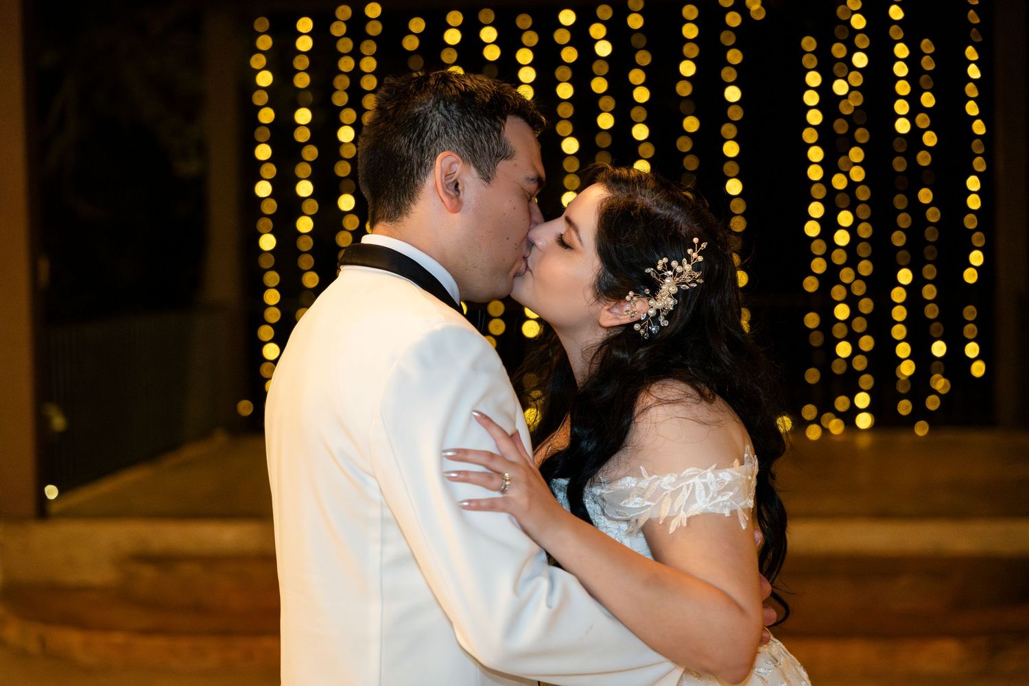 A romantic wedding couple shares a kiss in front of twinkling string lights at their evening reception.
