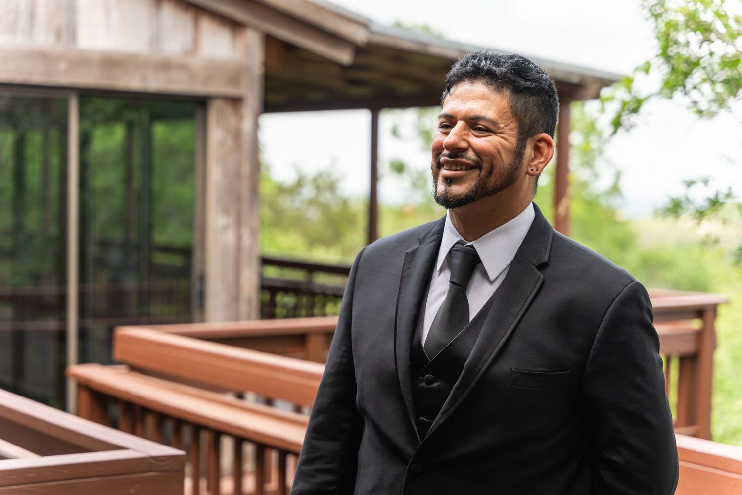 A person in formal attire smiles outdoors on a wooden deck wearing a black suit and tie.